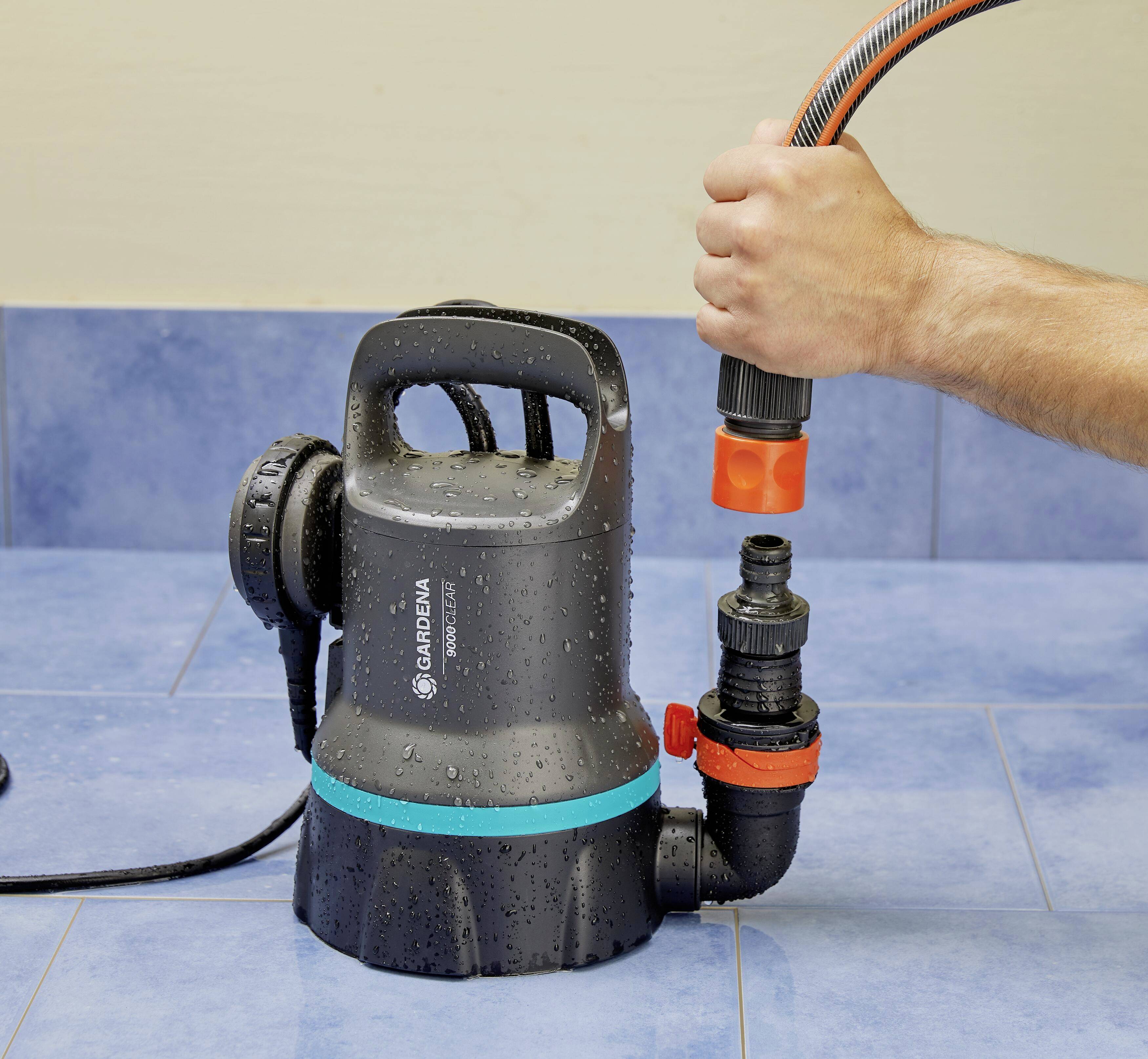 Een hand sluit een slang aan op een zwarte waterpomp die op een blauwe vloer staat. De pomp is nat van het water.