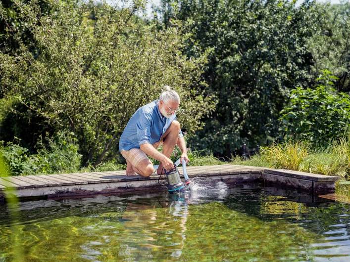 Een persoon knielt aan de rand van een vijver in een tuin en gebruikt een waterpomp om de vijver te vullen. Op de achtergrond zijn bomen en struiken zichtbaar.