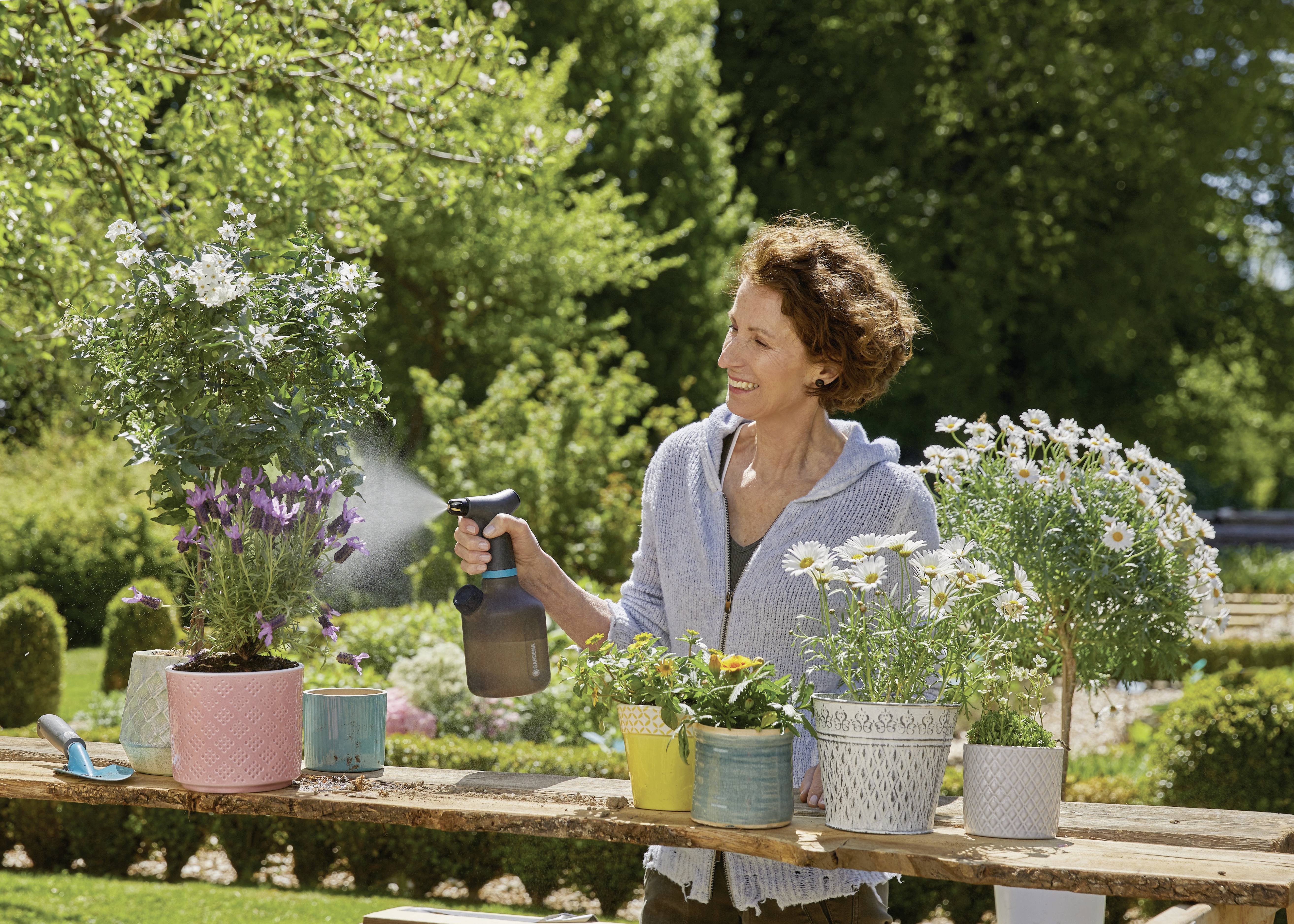 Een glimlachende vrouw besproeit bloemen met een spuitfles in de tuin. Verschillende potplanten staan op een houten tafel buiten.