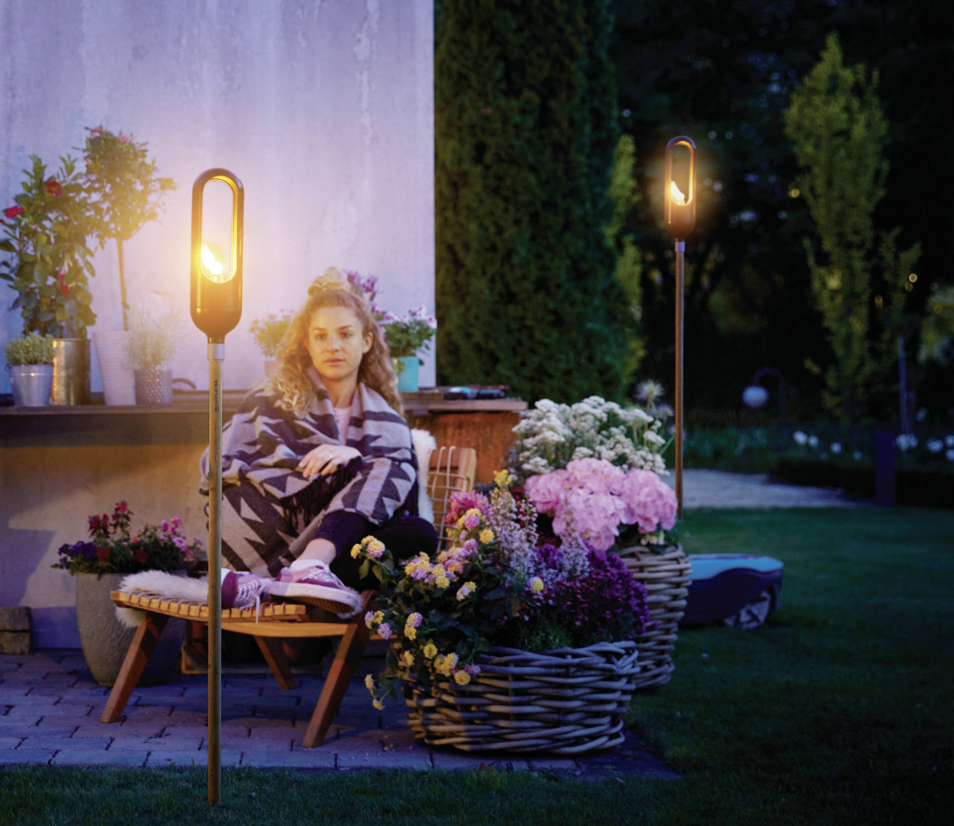 Een vrouw zit op een stoel in de tuin, omringd door bloemen en verlichte tuinlampen, bij het vallen van de avond.