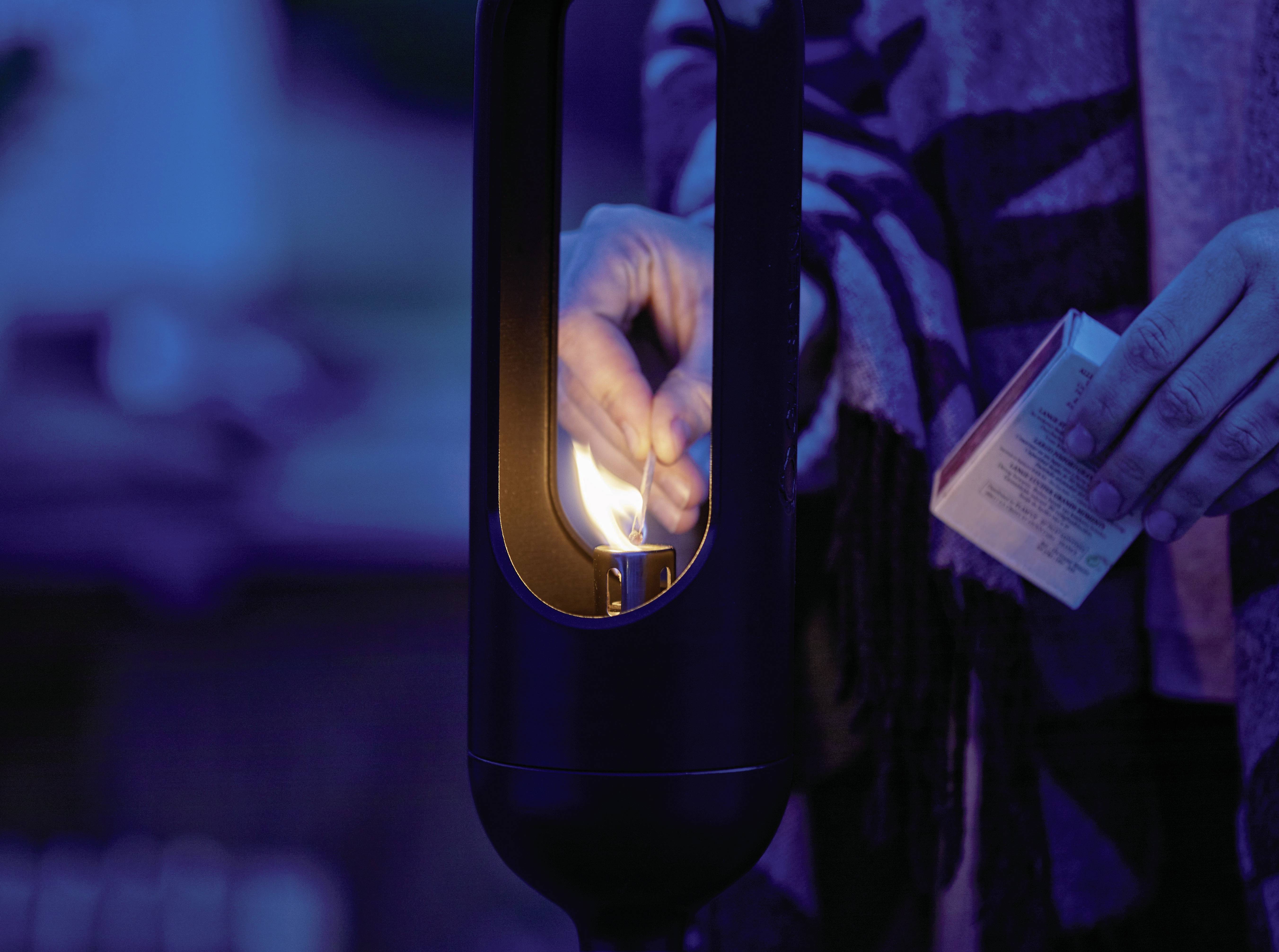 Iemand steekt een kaars aan in een donkere ruimte. De scène oogt rustig en sfeervol met warme verlichting.