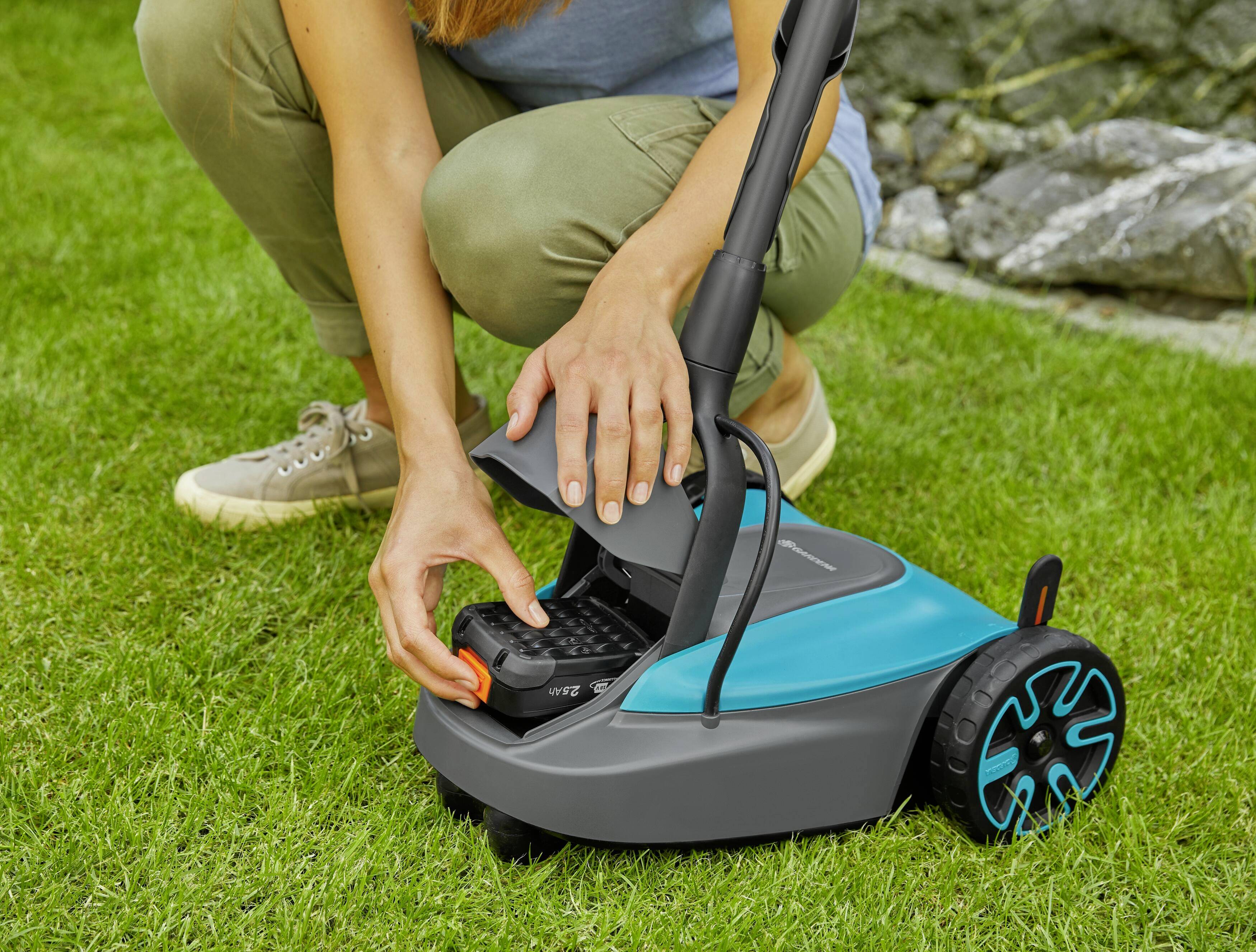 Een persoon vervangt de batterij van een grasmaaier. De grasmaaier is blauw en zwart, en de persoon draagt vrijetijdskleding.