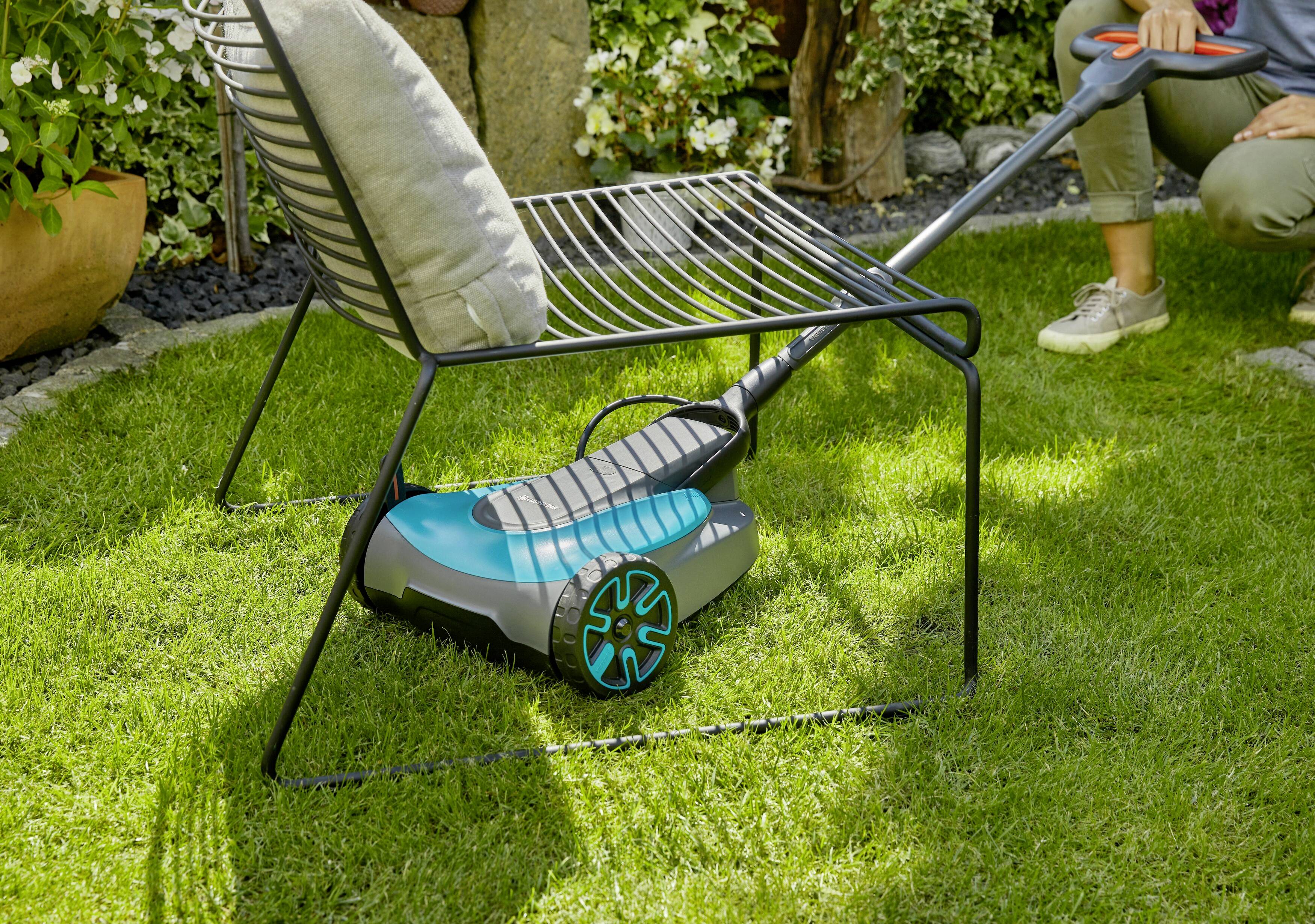 Een persoon maait het gazon in een tuin met een elektrische grasmaaier. Op de achtergrond staan planten en een stoel.