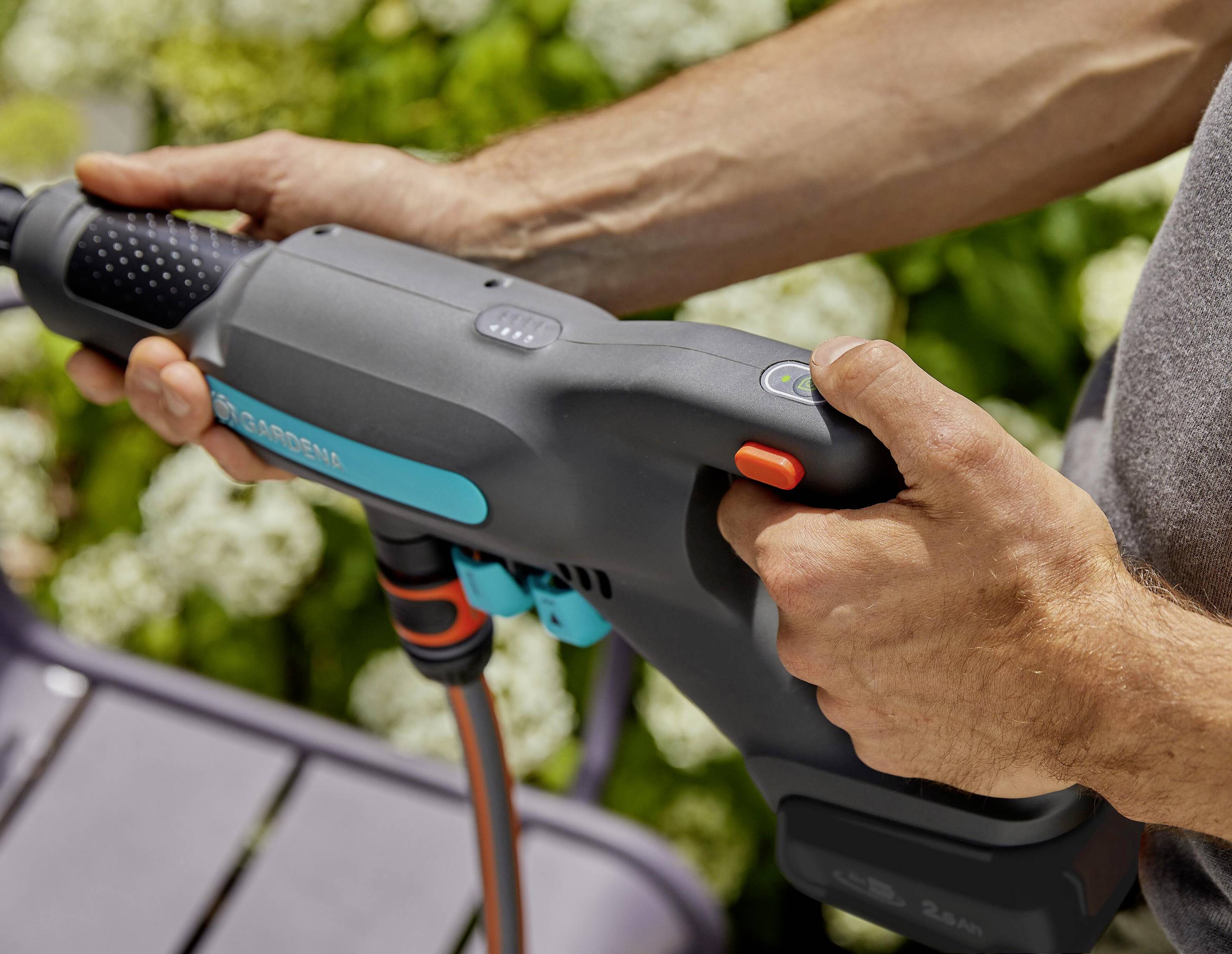 Een persoon houdt een hogedrukreiniger in de handen, klaar voor gebruik in de tuin. Op de achtergrond zijn groene planten zichtbaar.