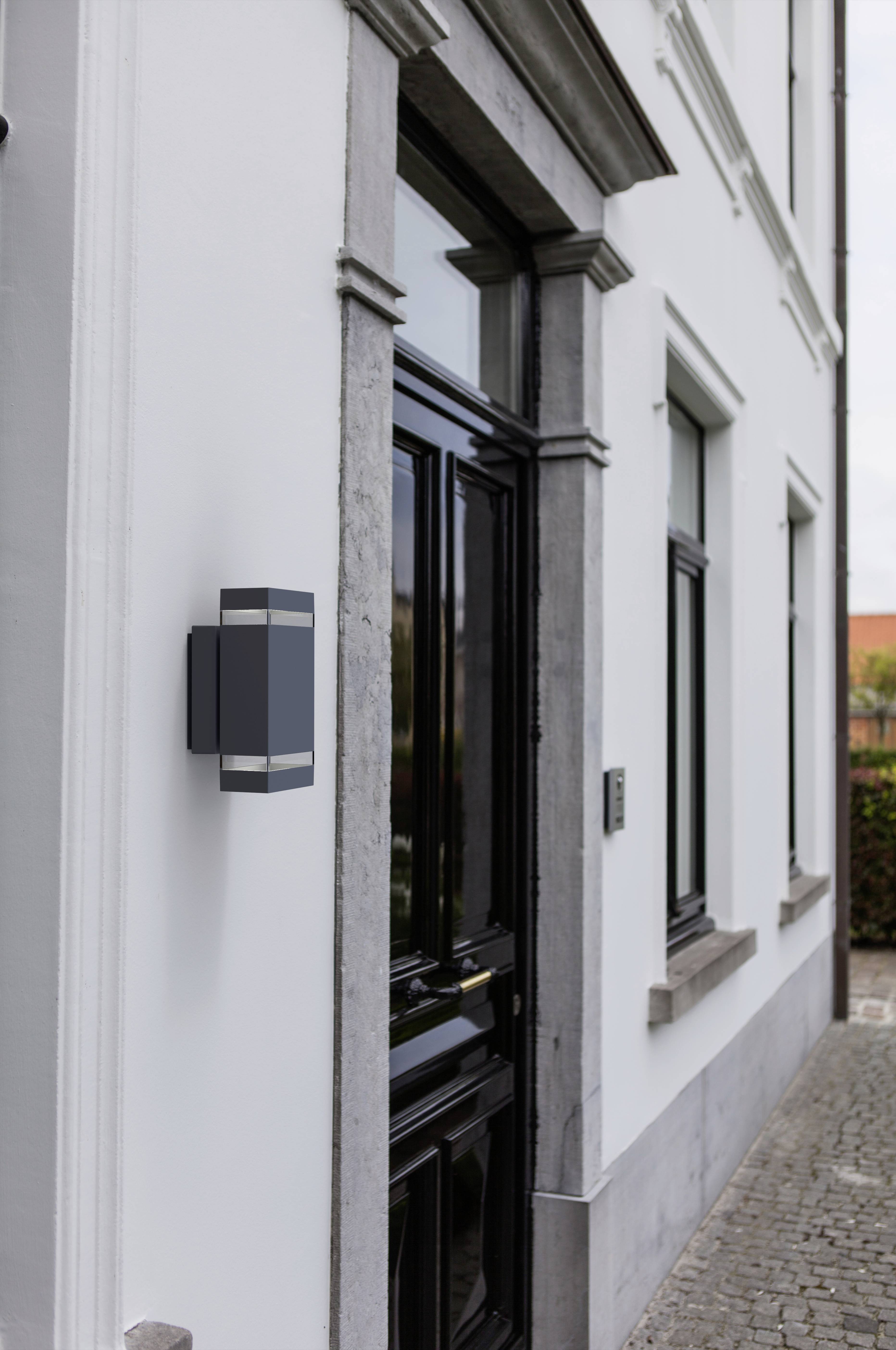 Huisgevel met zwarte houten deur en moderne buitenverlichting. Links naast de deur bevindt zich een deurbel. Een pad van keien leidt naar de deur.