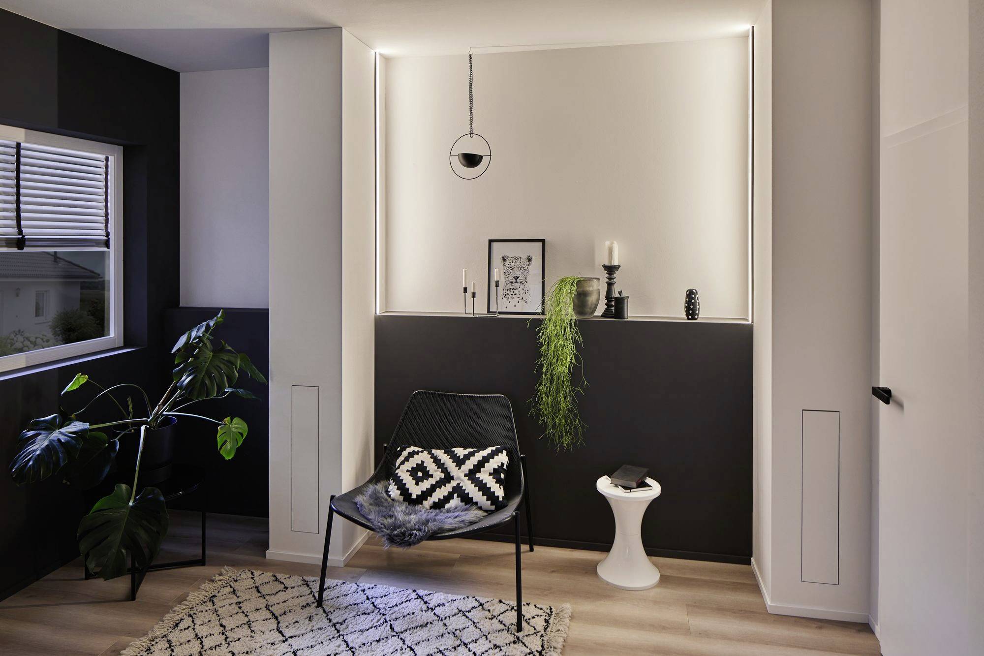 Een minimalistisch woonkamer met een zwarte stoel, kussens, vloerkleed, planten, kaars en kunstwerk op een rek.