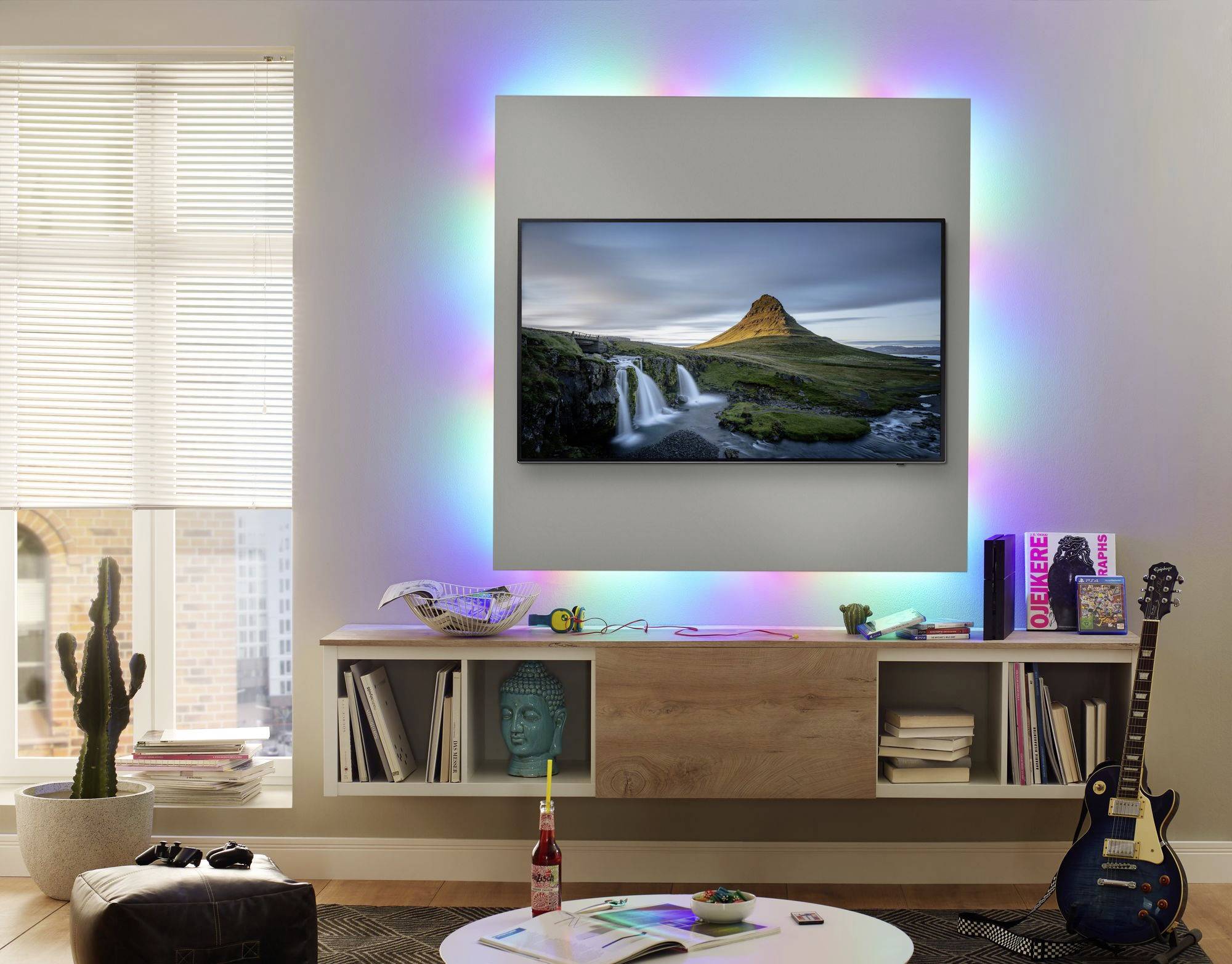 Woonkamer met aan de muur gemonteerde televisie die een landschap toont. Omgeven door kleurrijke LED-verlichting, met decoratieve items op de planken eronder.