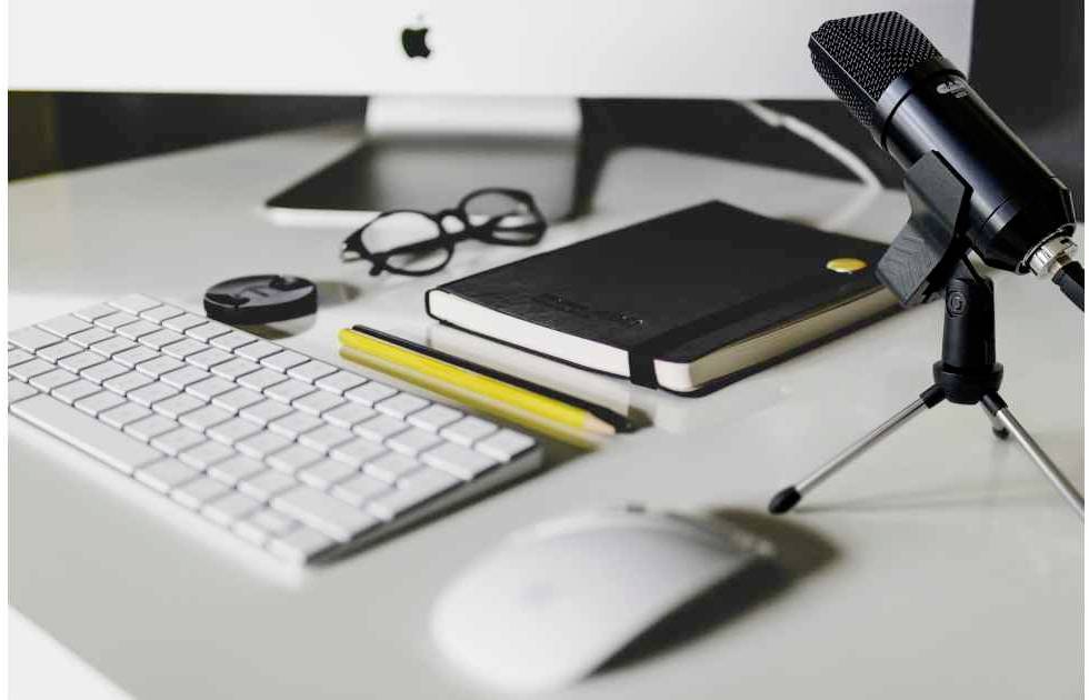 Bureau met computerkeyboard, muis, notitieboek, bril, microfoon en potlood, voor een computerscherm.