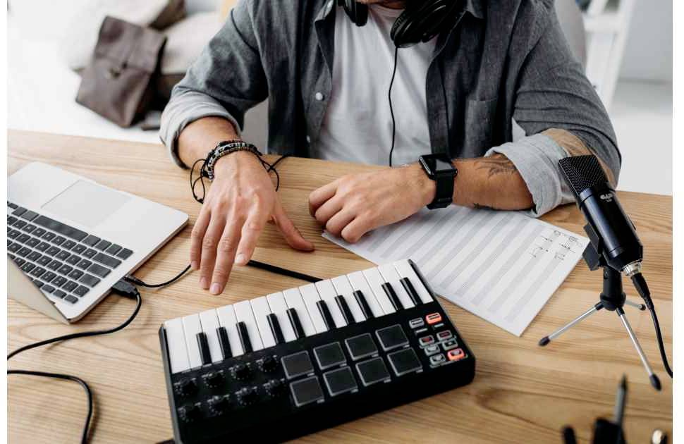 Een persoon aan een bureau met een laptop, een MIDI-keyboard, bladmuziek en een microfoon, die muziek componeert of opneemt.