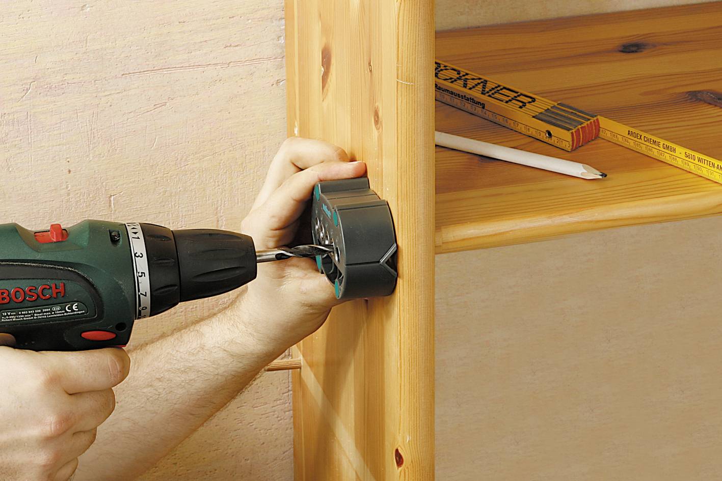 Een persoon boort met een boormachine een gat in een houten plank. Een potlood en een meetlat liggen op de plank.