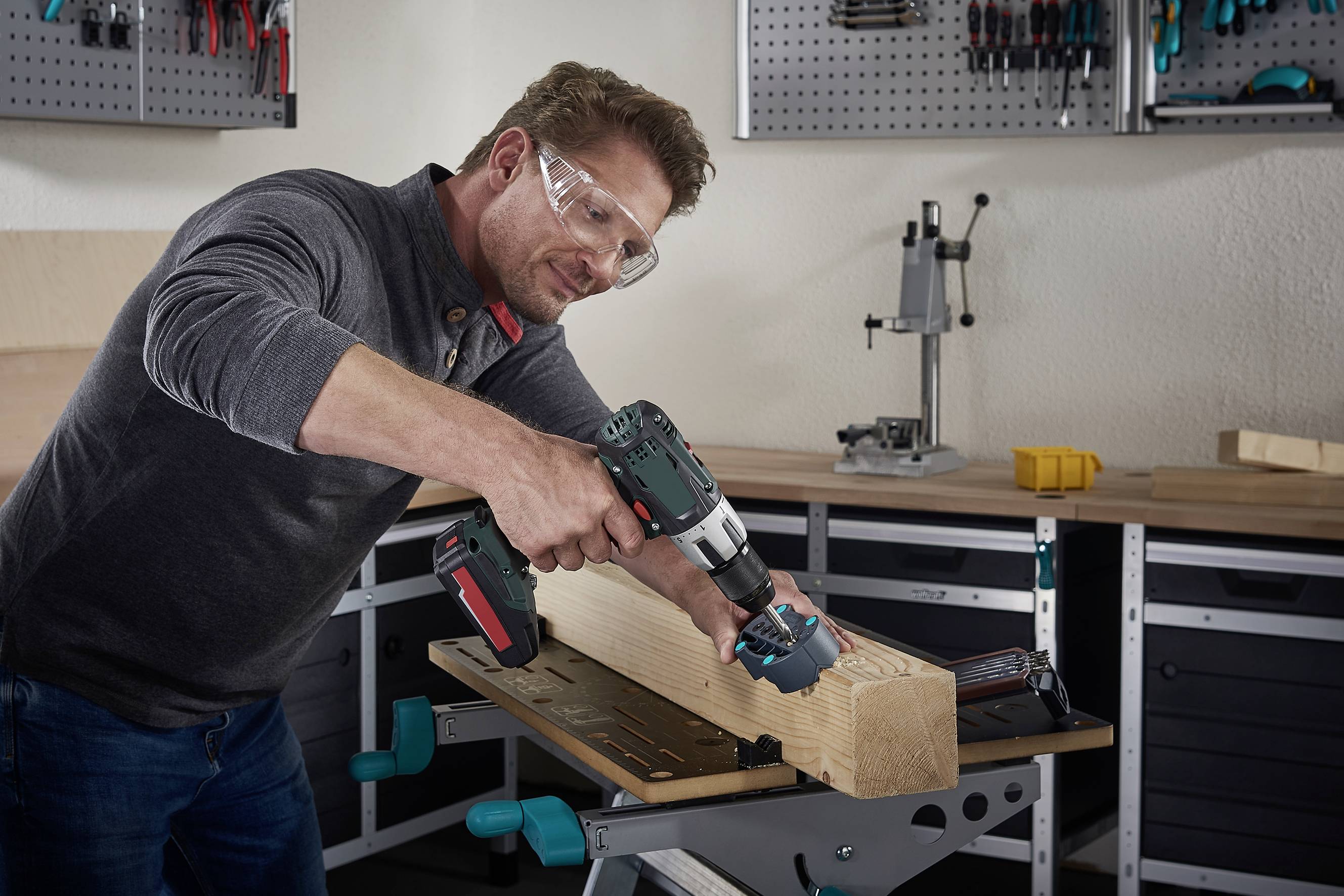 Een man boort een gat in een houten blok met een elektrische boormachine, terwijl hij een veiligheidsbril draagt. Gereedschappen op de achtergrond.