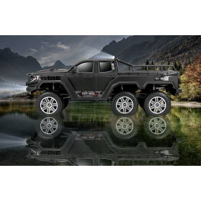 Een grote, zwarte terreinwagen met zes wielen staat aan de oever van een meer, met een gespiegelde weerspiegeling in het water en nevelige bergen op de achtergrond.