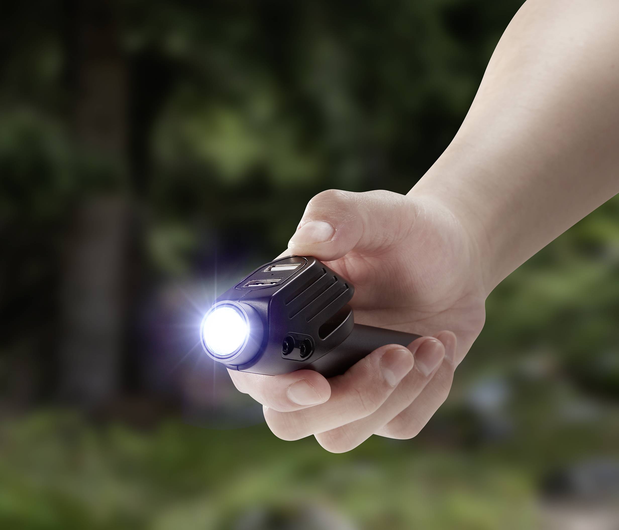 Een hand houdt een felle zaklamp gericht op een bos. De achtergrond is wazig met bomen.