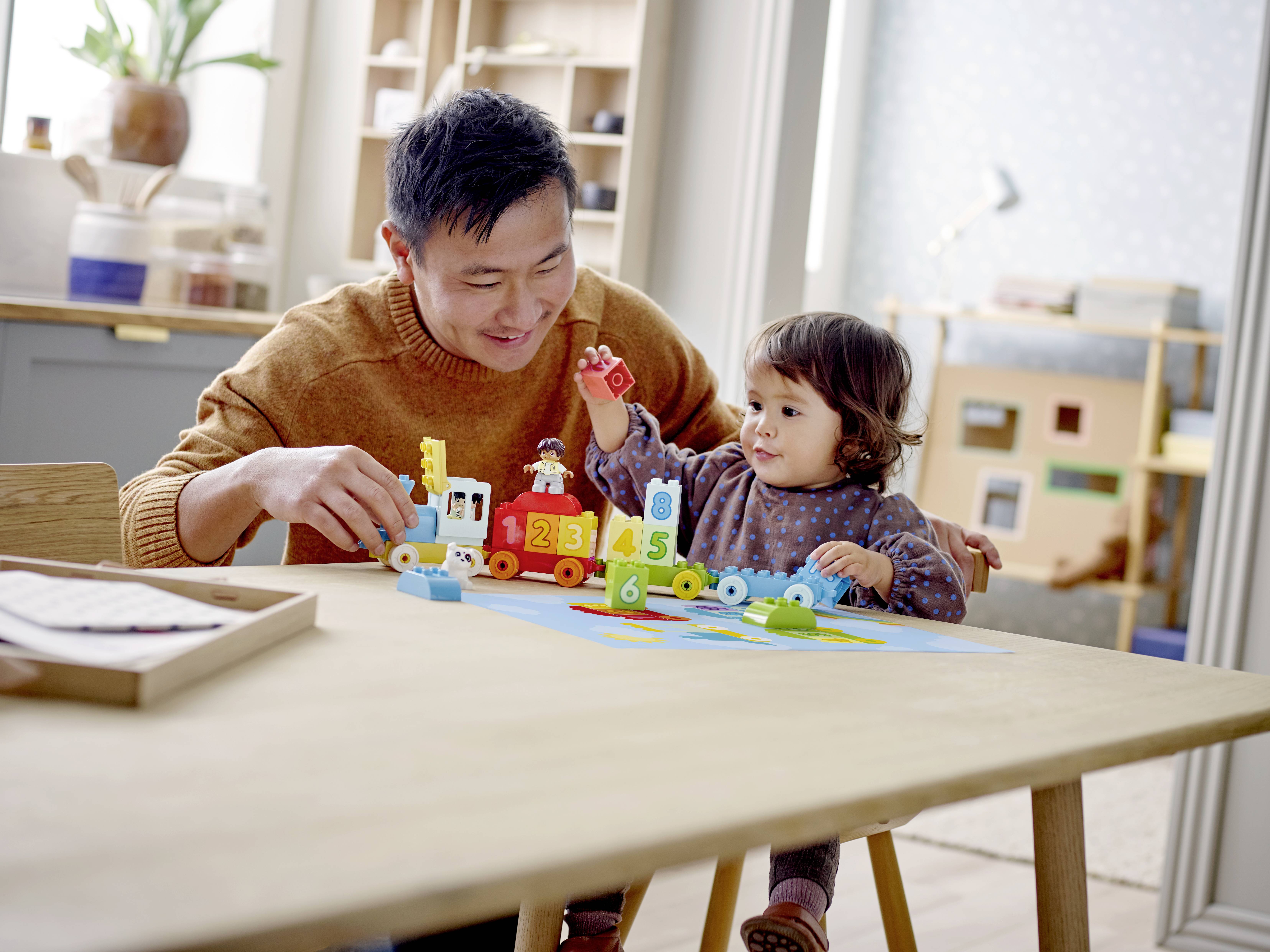 Een volwassene en een peuter spelen met kleurrijke bouwblokken aan een tafel in een woonkamer. De volwassene lacht.