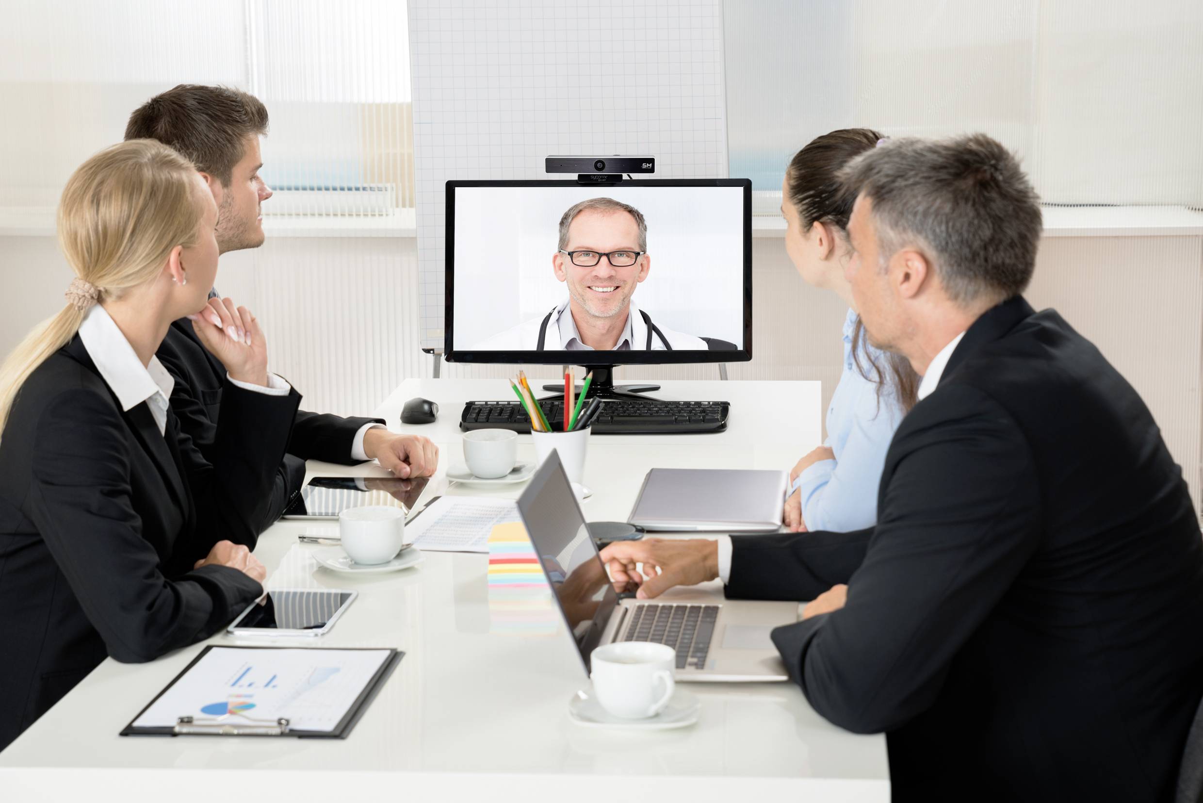 Vijf personen nemen deel aan een videoconferentie in een vergaderzaal op kantoor. Op het scherm is een lachende man te zien.