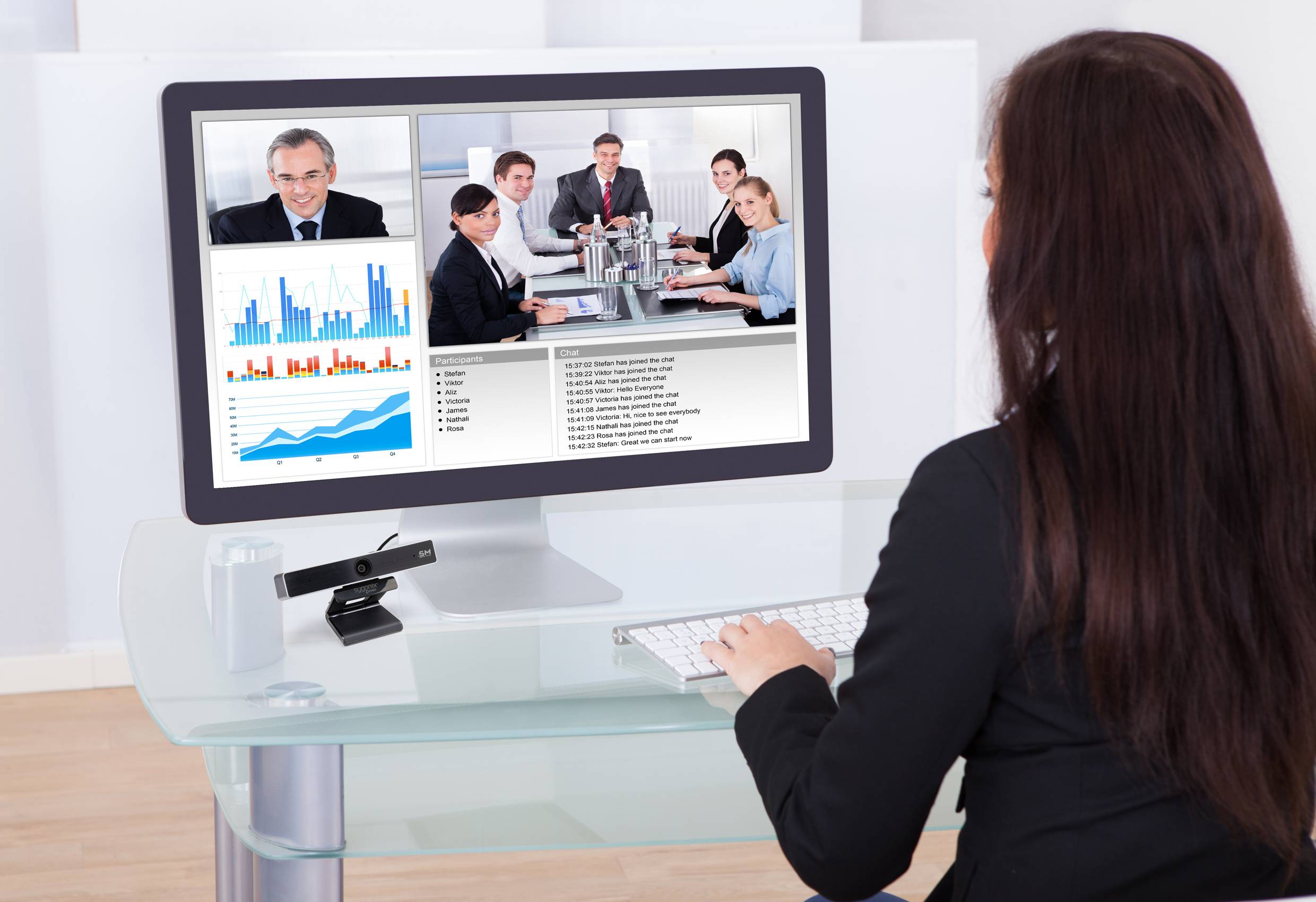 Een vrouw zit aan een tafel en neemt deel aan een videoconferentie. Op het scherm zijn diagrammen en meerdere conferentiedeelnemers te zien.
