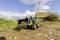 Een groene tractor met voorlader vervoert grond op een landelijke veldweg, met op de achtergrond een oude schuur.