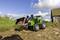 Een groene speelgoedtractor met een schep vervoert aarde over een veldweg. Op de achtergrond is een schuur te zien.
