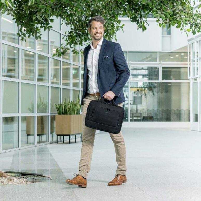 Een man in een pak loopt door een modern kantoorgebouw. Hij draagt een laptoptas en glimlacht terwijl hij langs een groot raam loopt.
