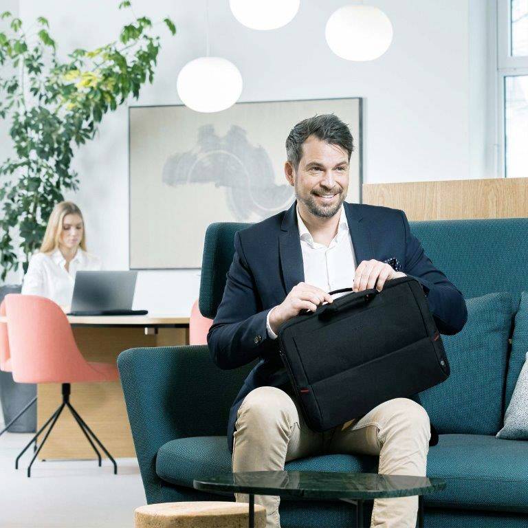 Een man in een pak zit op een bank en houdt een tas vast; op de achtergrond werkt een vrouw achter een laptop aan een bureau.