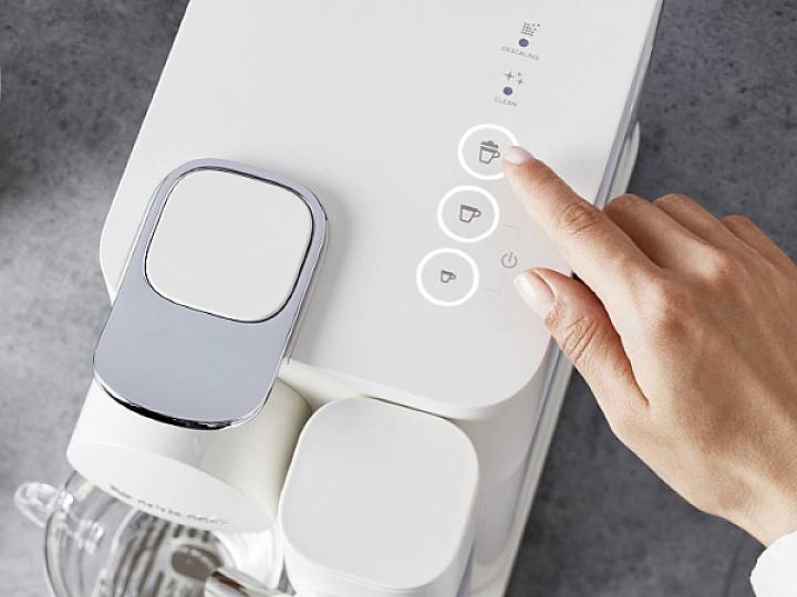 Hand drukt knop op witte koffiemachine. Functies: Espresso, Cappuccino, Reiniging. Machine op grijze oppervlakte.