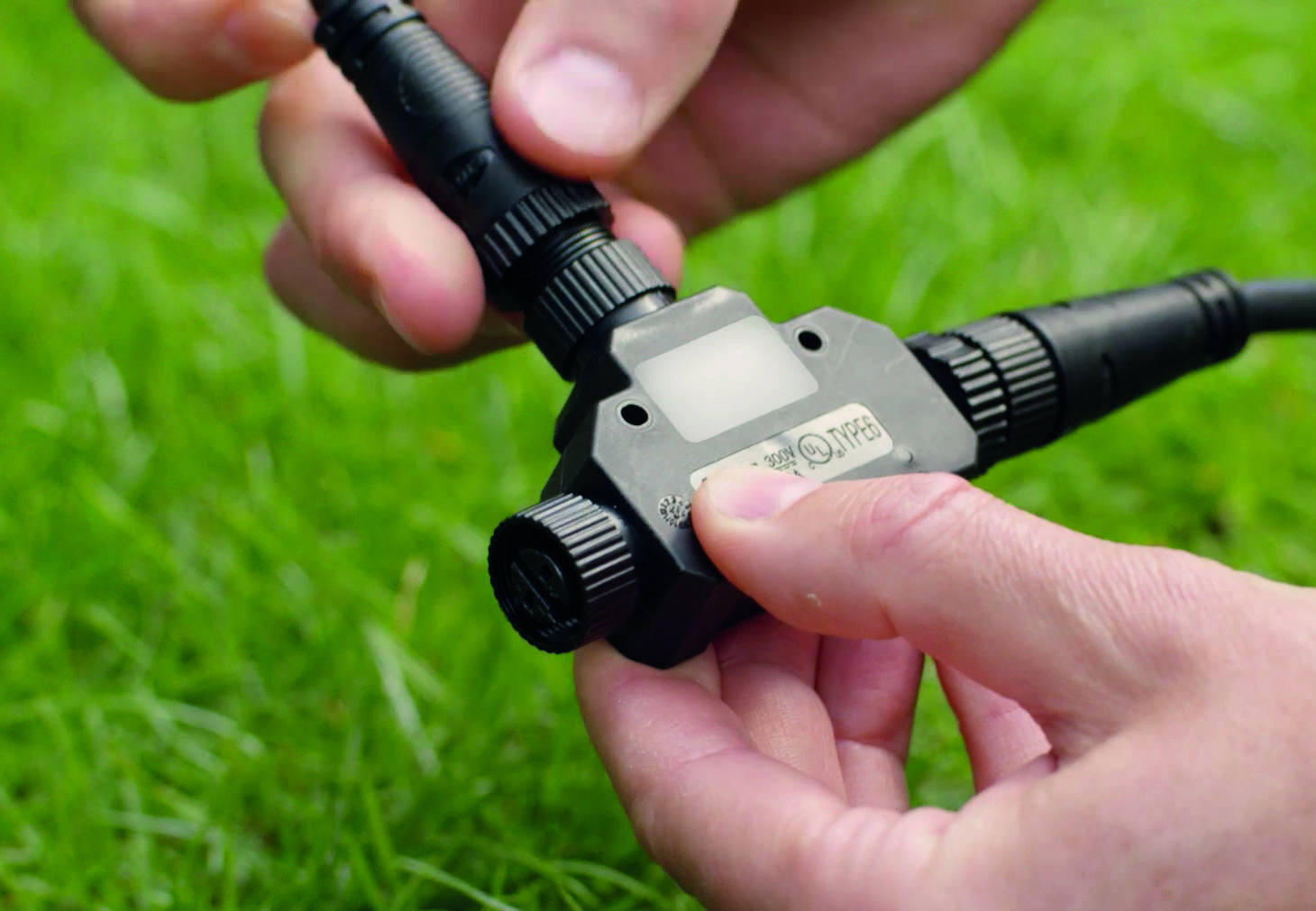 Een persoon houdt een zwarte kabel met aansluitingen in de handen. De achtergrond toont groen gras.