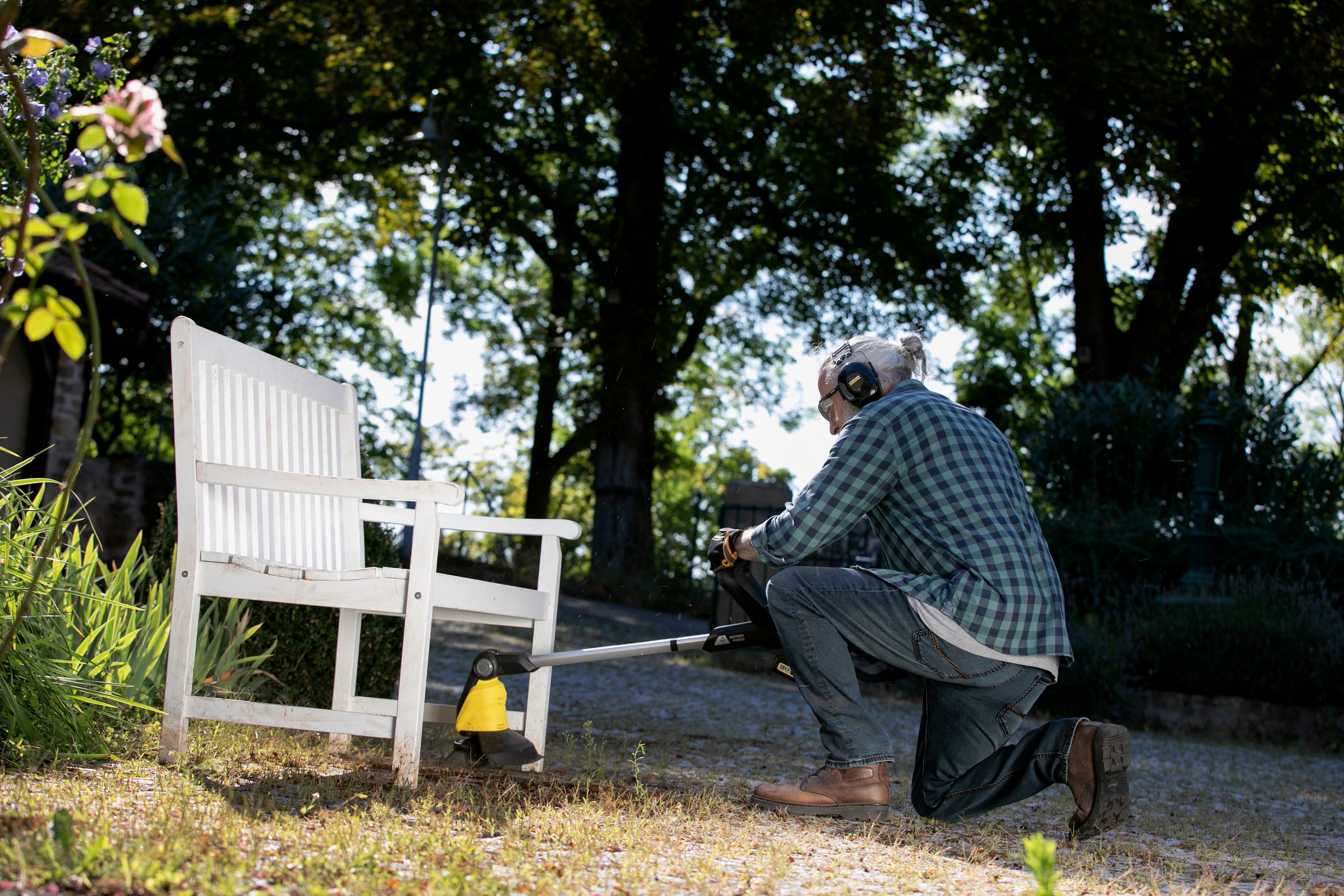 Een persoon in beschermende kleding knielt in de tuin en maait gras rond een witte bank met een bosmaaier.