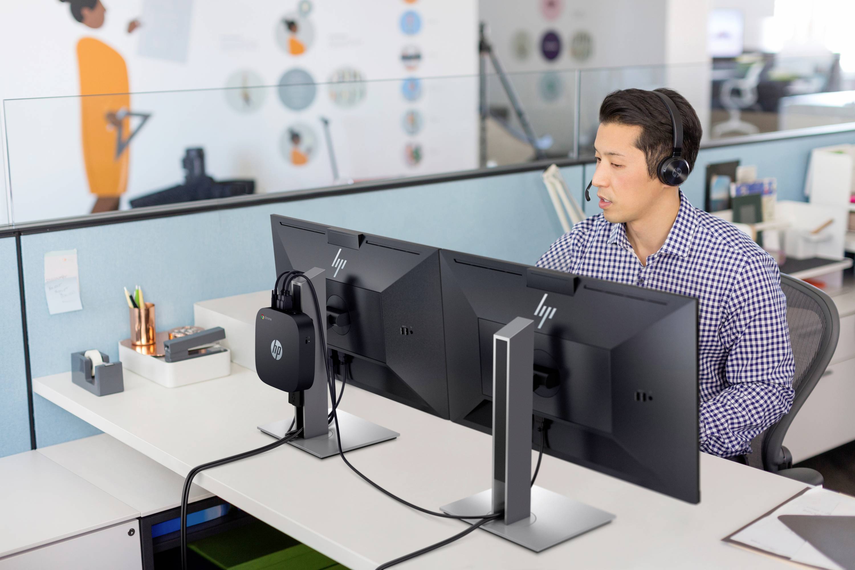 Een man met koptelefoon zit in een modern kantoor voor twee monitors, geconcentreerd aan het werk, met kantoorbenodigdheden op de achtergrond.