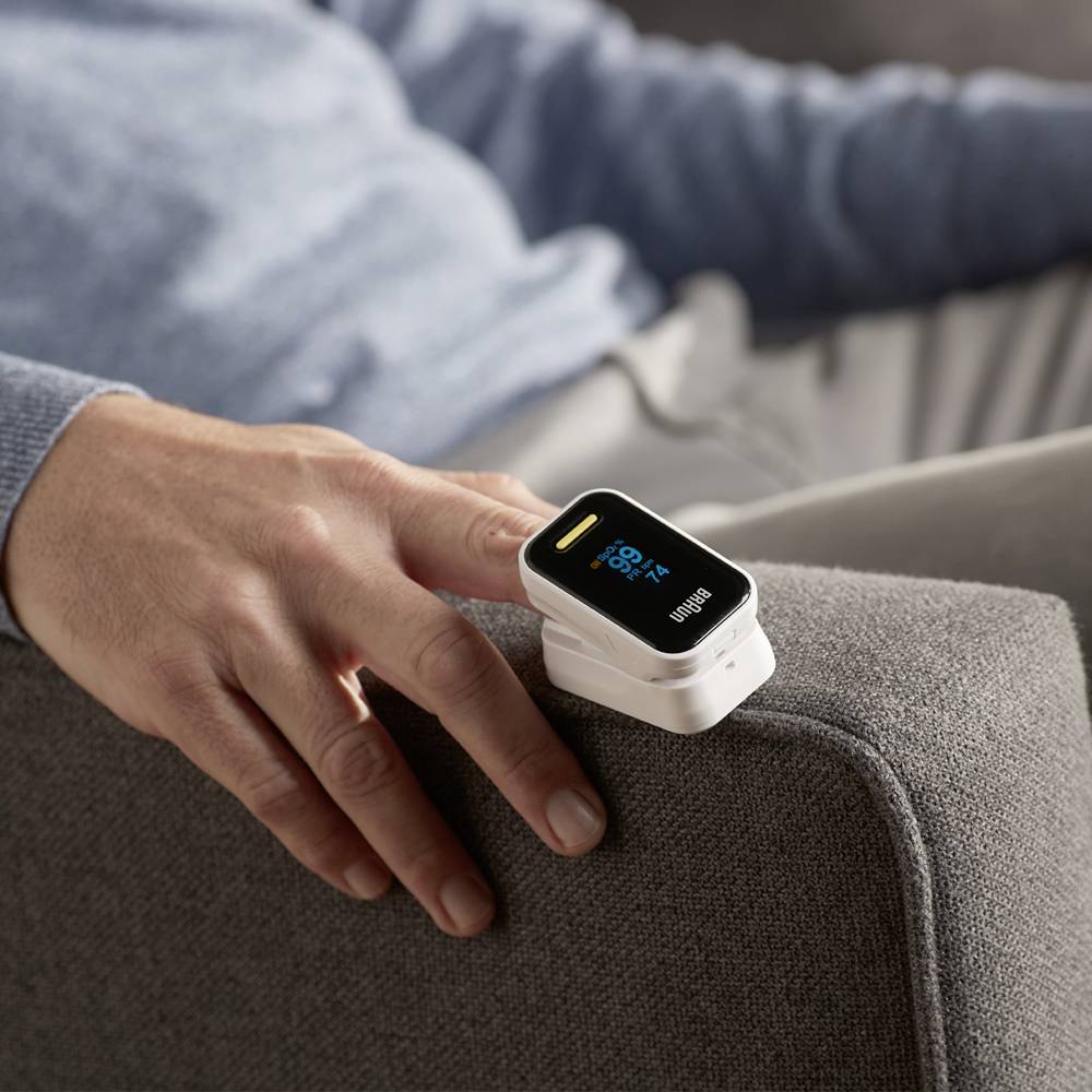 Een persoon meet zijn zuurstofgehalte met een pulsoxymeter aan de vinger. Het apparaat geeft 98% aan. De persoon zit ontspannen.
