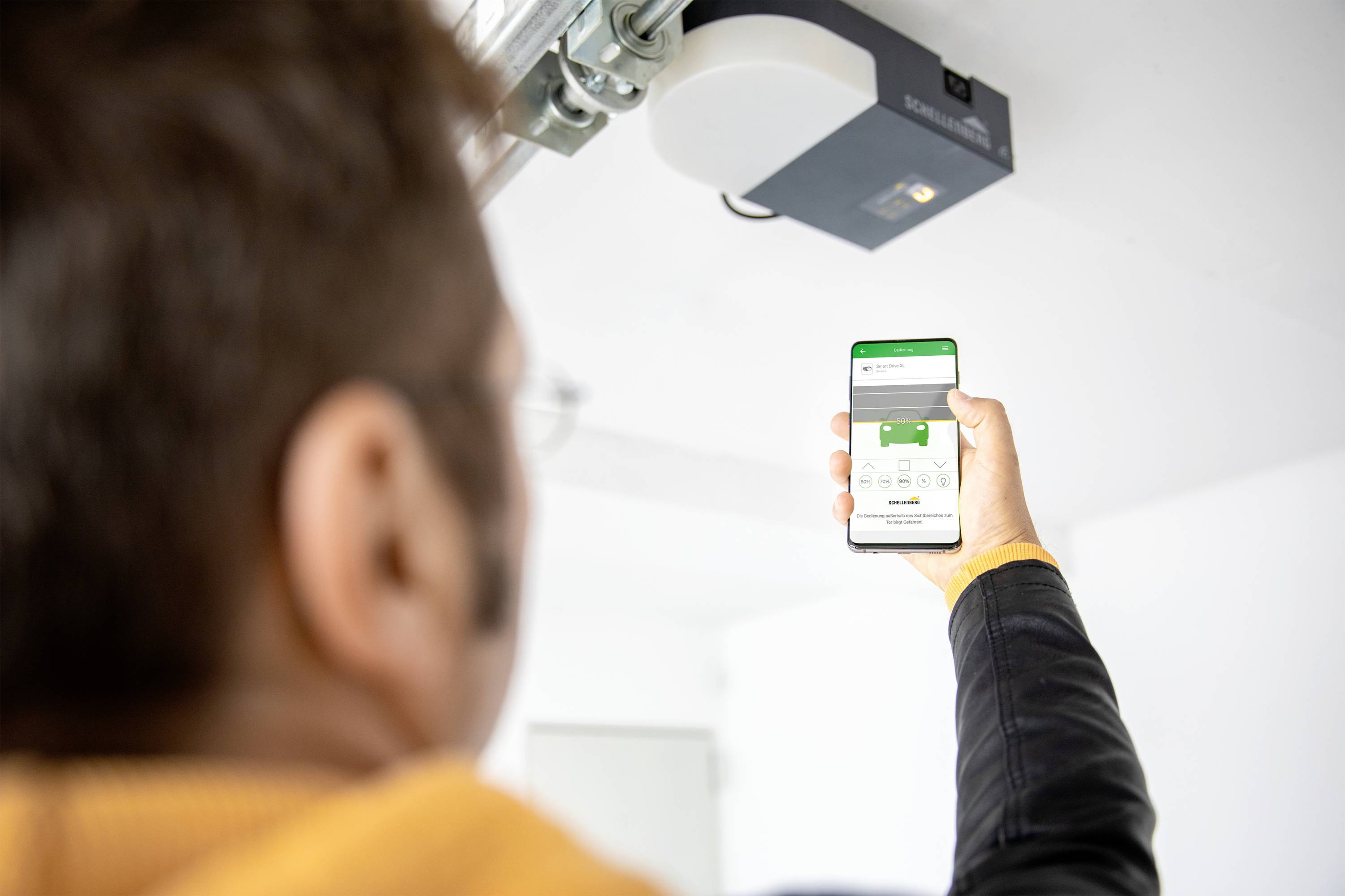 Een man bedient een garagedeur met zijn smartphone. Op het scherm is een app te zien waarmee de poort wordt bediend.