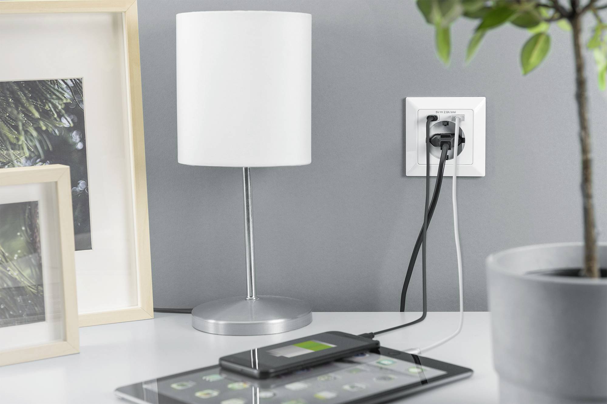 Een tafellamp, een tablet en een smartphone worden opgeladen. De opladers zijn aangesloten op een stopcontact aan de muur.