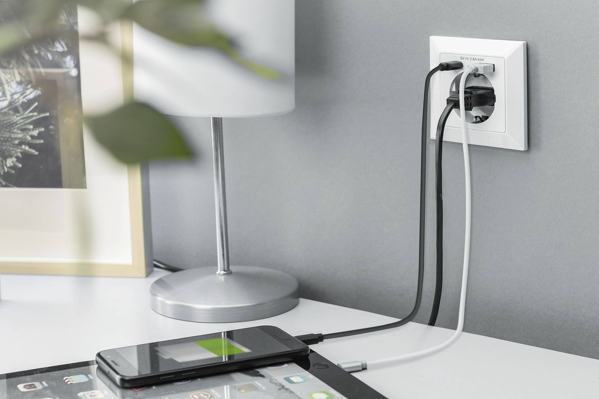 Smartphone en tablet liggen op een tafel, beide worden via kabels opgeladen aan een wandcontactdoos. Op de achtergrond een lamp.