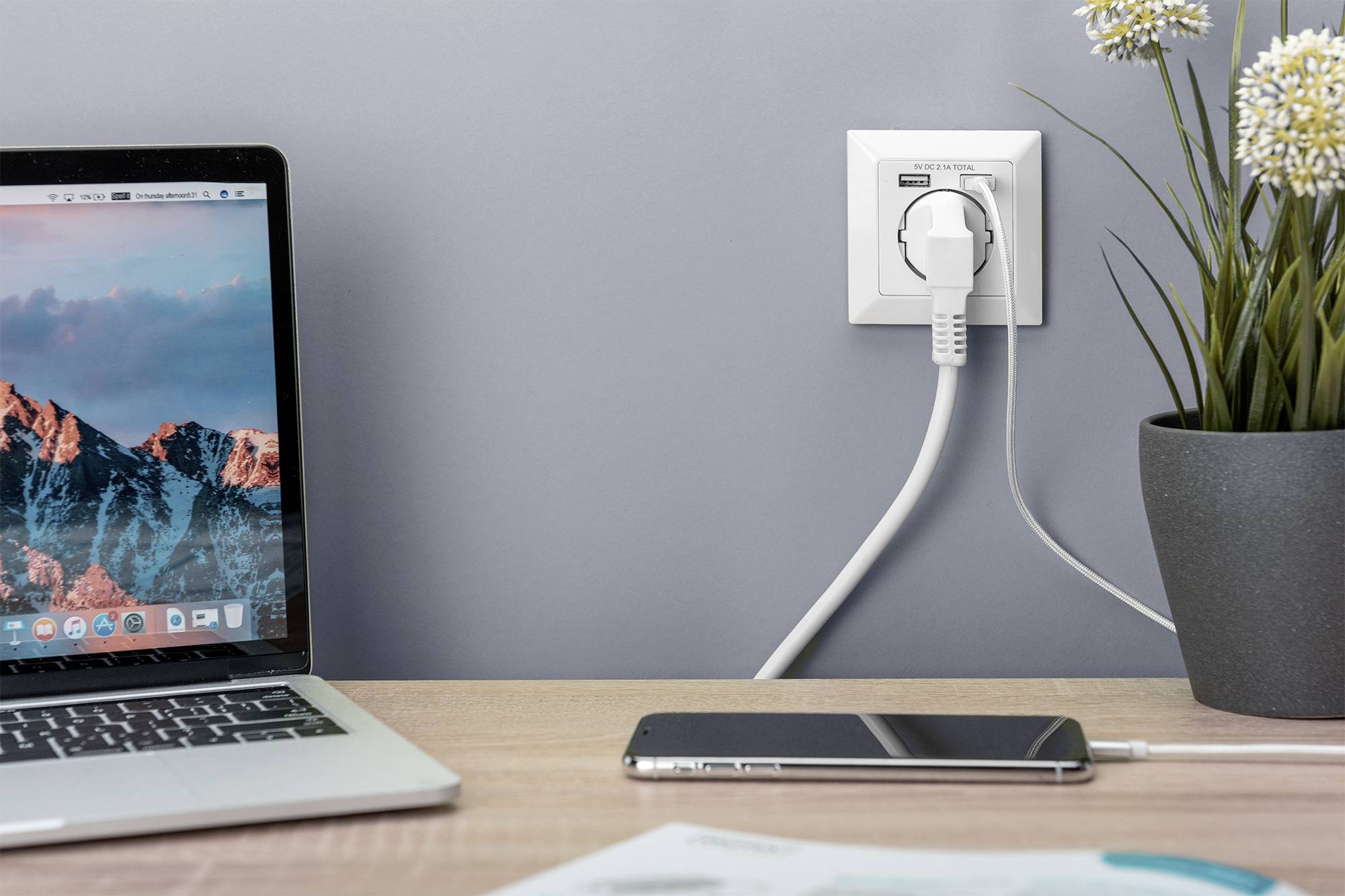 Een laptop, een smartphone en een stopcontact met aangesloten kabel op een tafel naast een plant.