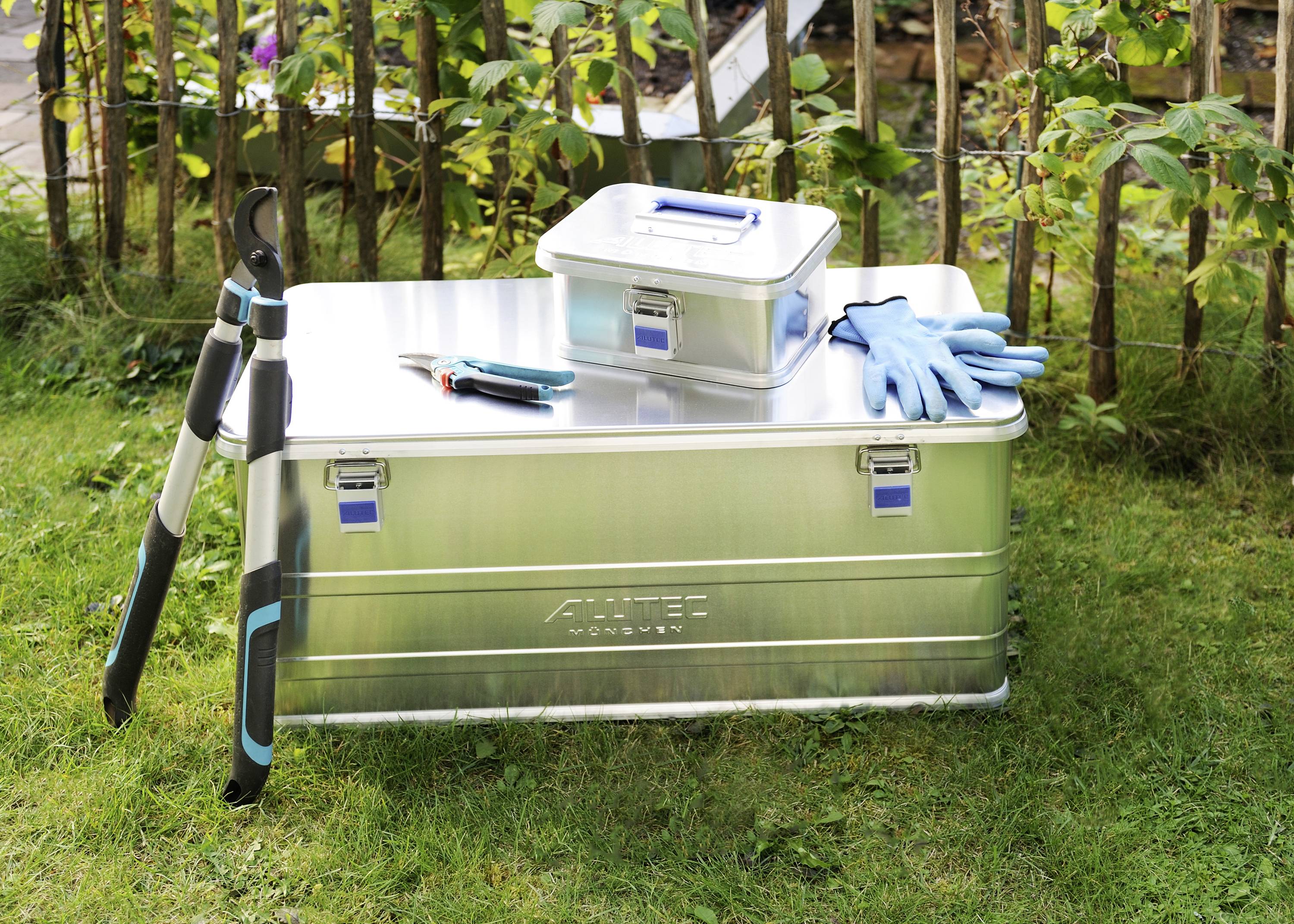 Grote, zilveren metalen kist op gras met een kleinere kist, tuinhandschoenen, takkenschaar en tuinschaar ernaast; tuin op de achtergrond.