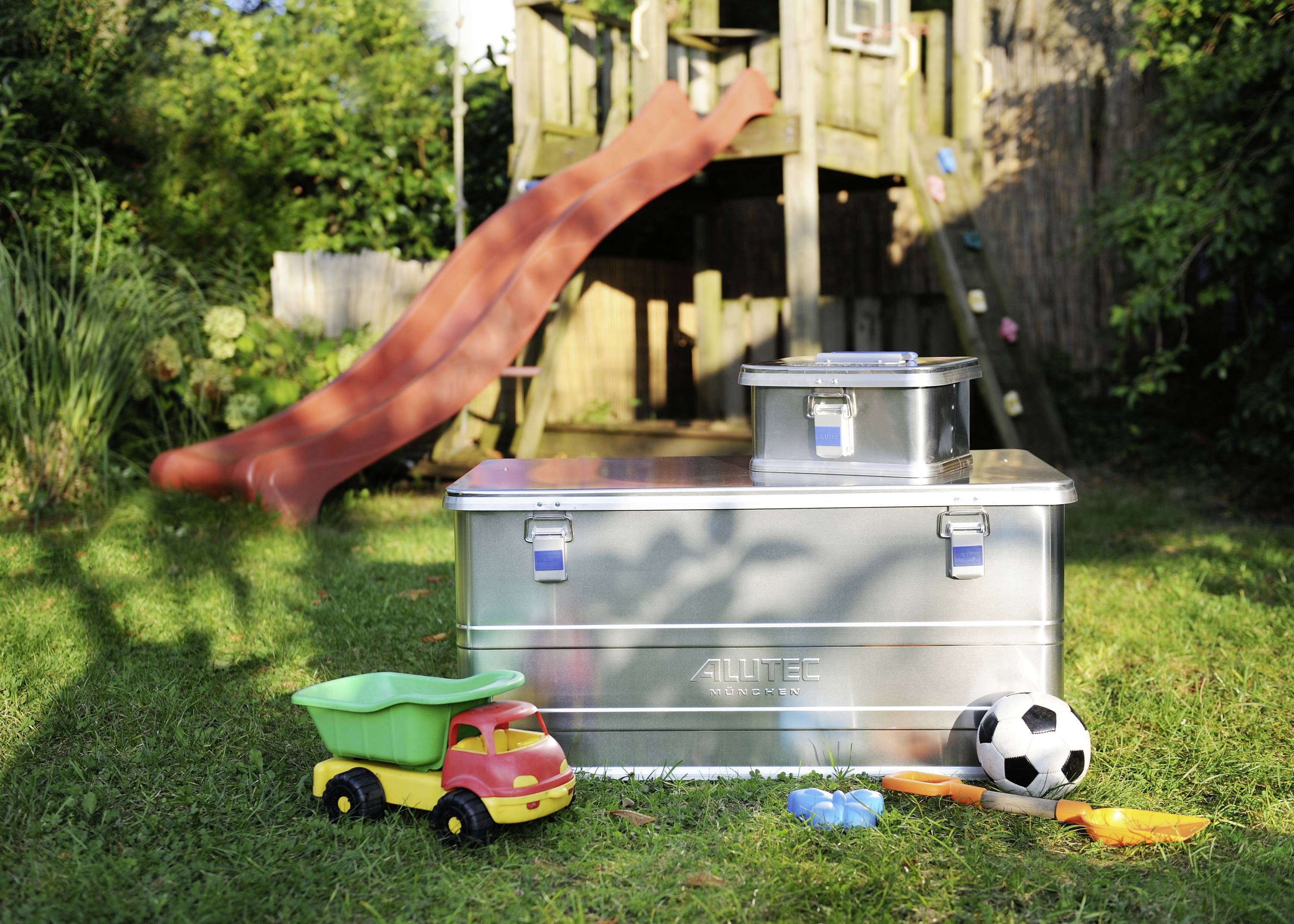 Metalen kisten op een grasveld voor een klimrek met glijbaan. Daarvoor liggen kleurrijke speelgoederen zoals een graafmachine en een voetbal.