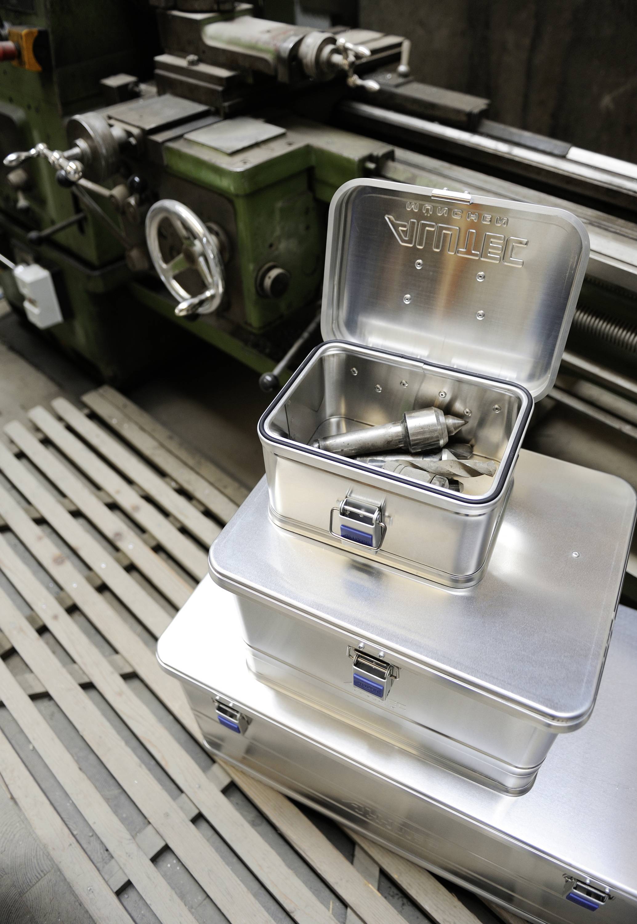 Drie metalen dozen op de voorgrond in een werkplaats; een is open met gereedschappen. Op de achtergrond een groene draaibank.