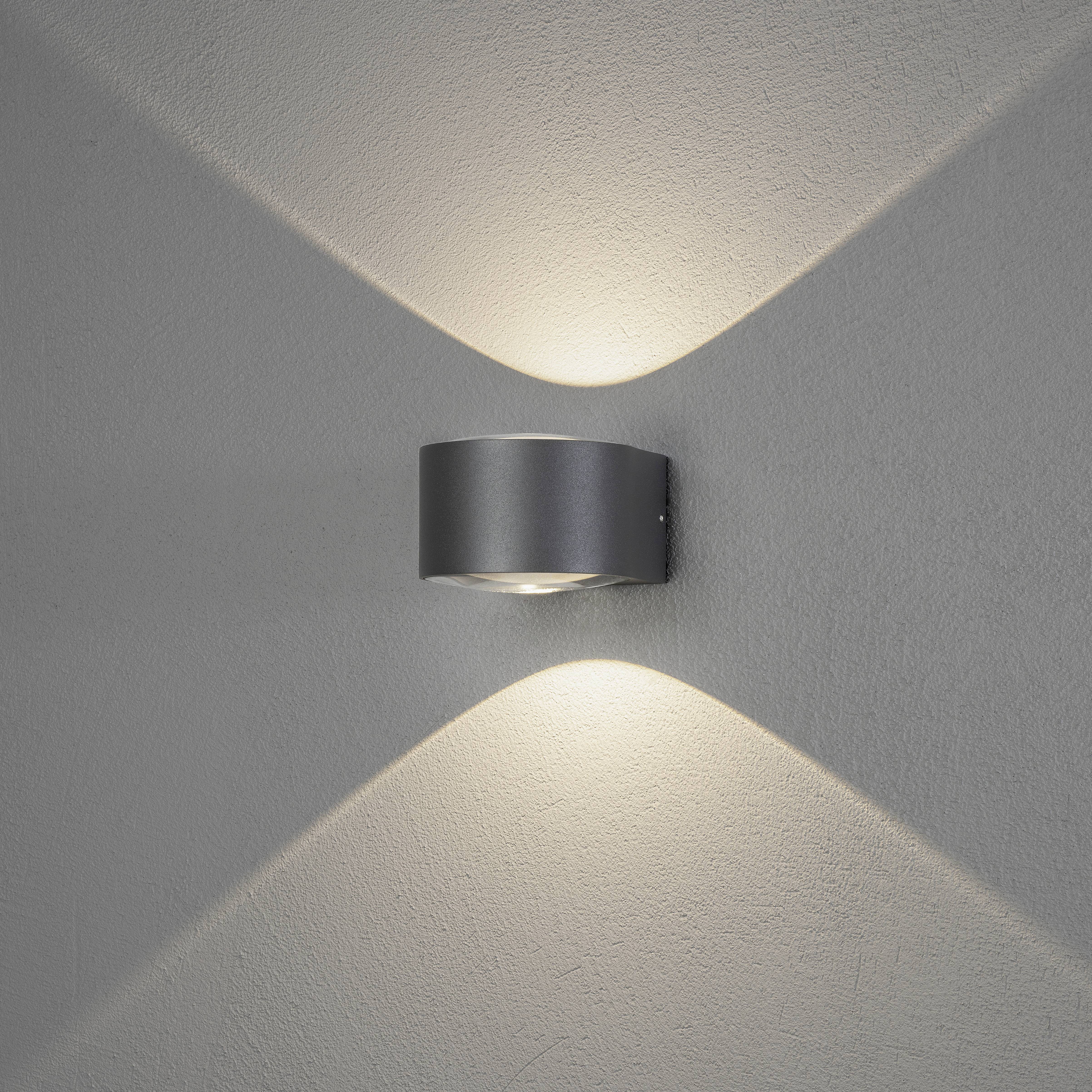 Een moderne wandlamp van metaal aan een grijze muur, die zacht licht uitstraalt naar boven en beneden en een X-patroon creëert.