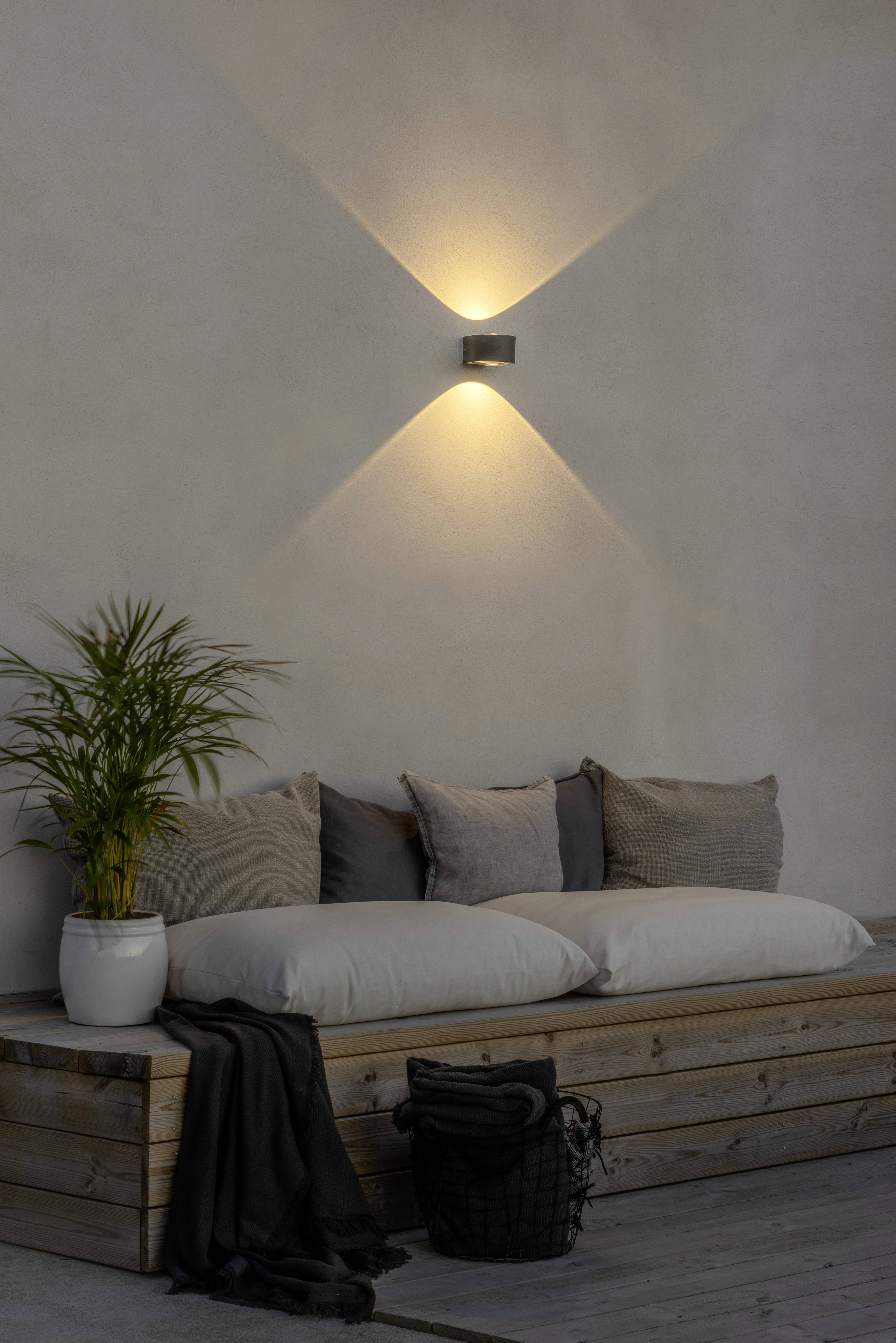 Een gezellige houten vloer met kussens en een deken erop, naast een potplant. Aan de muur erboven schijnt een wandlamp.