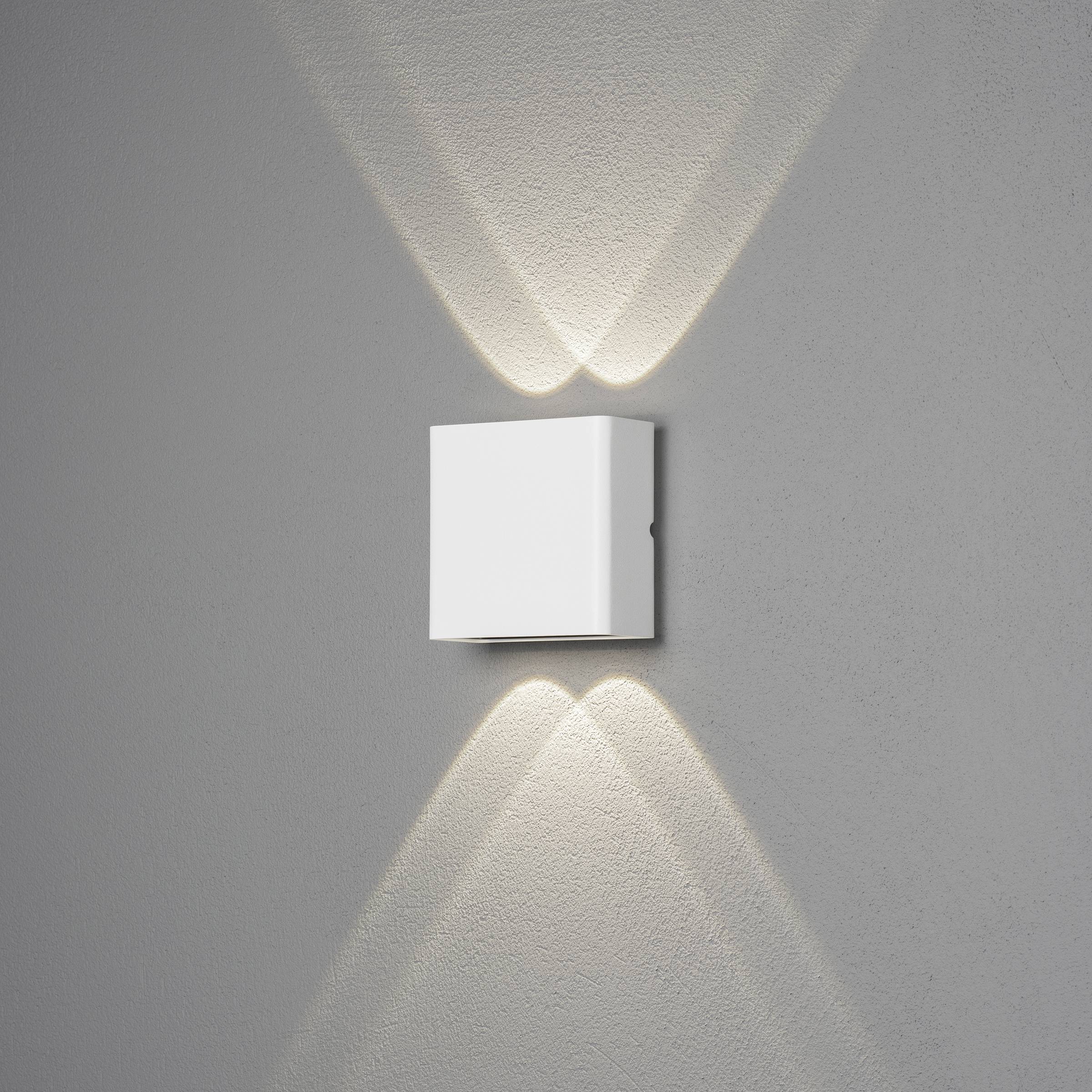 Witte, vierkante wandlamp straalt warm licht naar boven en beneden op grijze muur, creëert V-patroon.