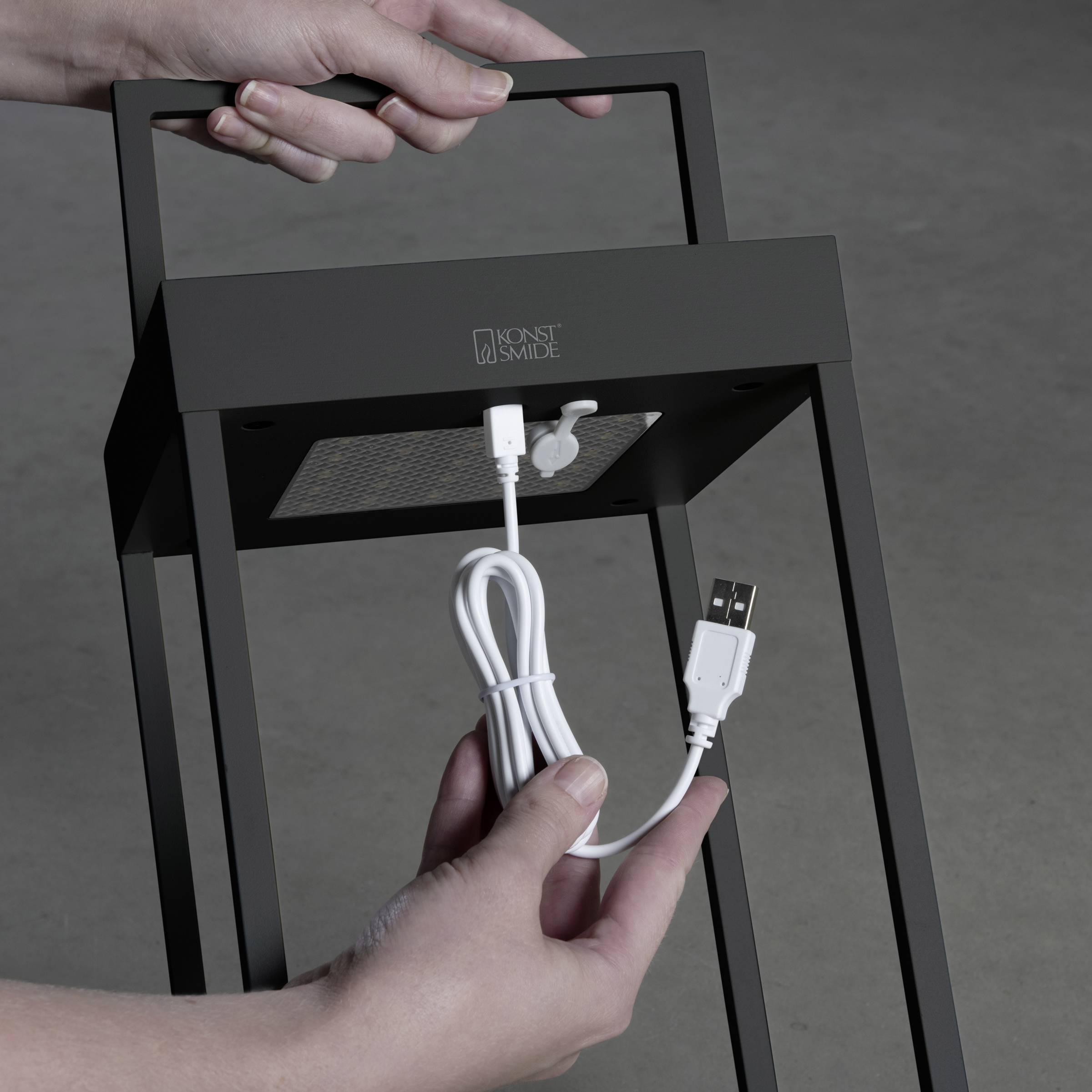 Een persoon houdt een oplaadkabel vast die wordt aangesloten op de USB-poort van een tafellamp. De lamp heeft een moderne vormgeving.