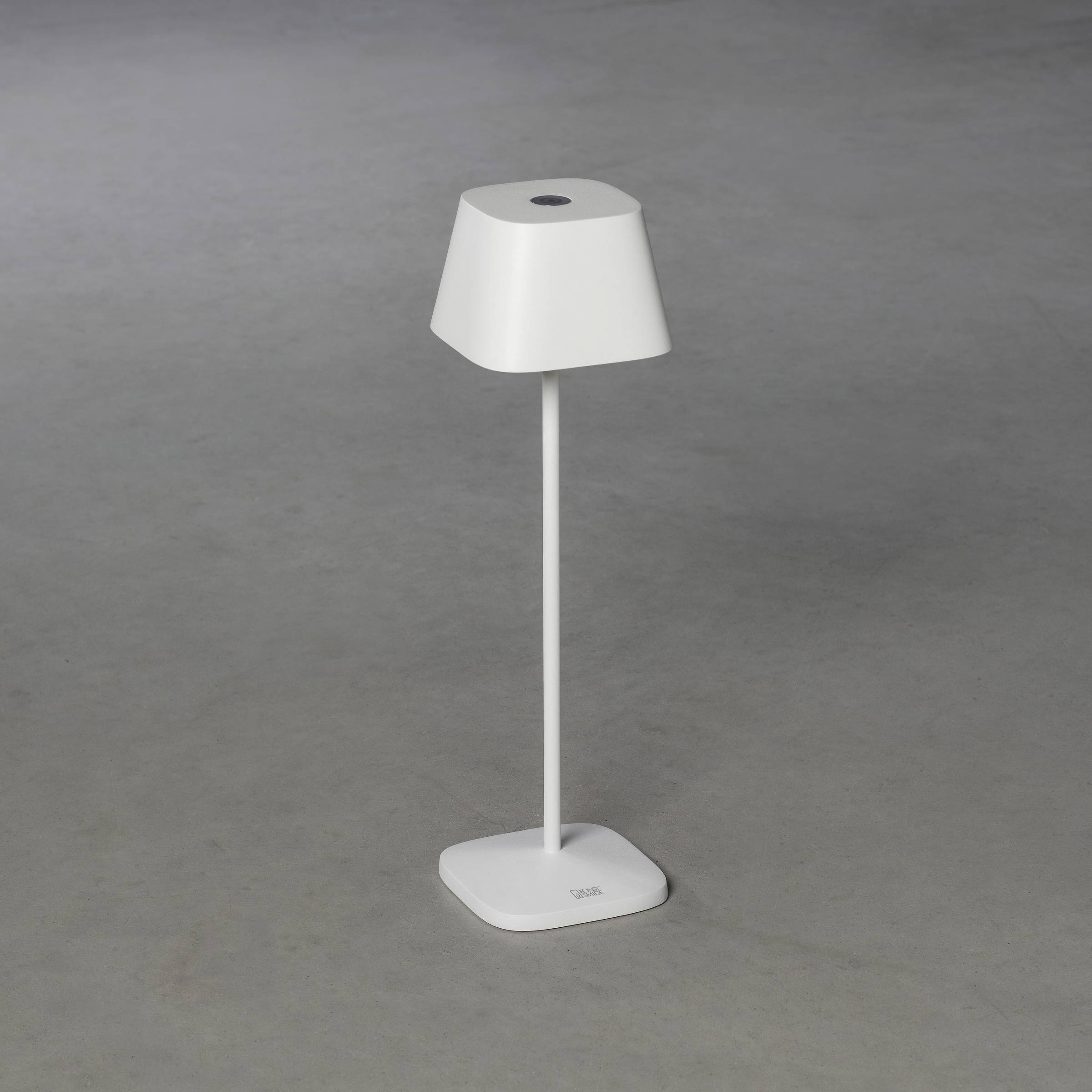 Witte tafellamp met een modern, minimalistisch ontwerp op een grijze achtergrond. Vierkante voet, slanke poot, trapezevormige lampenkap.