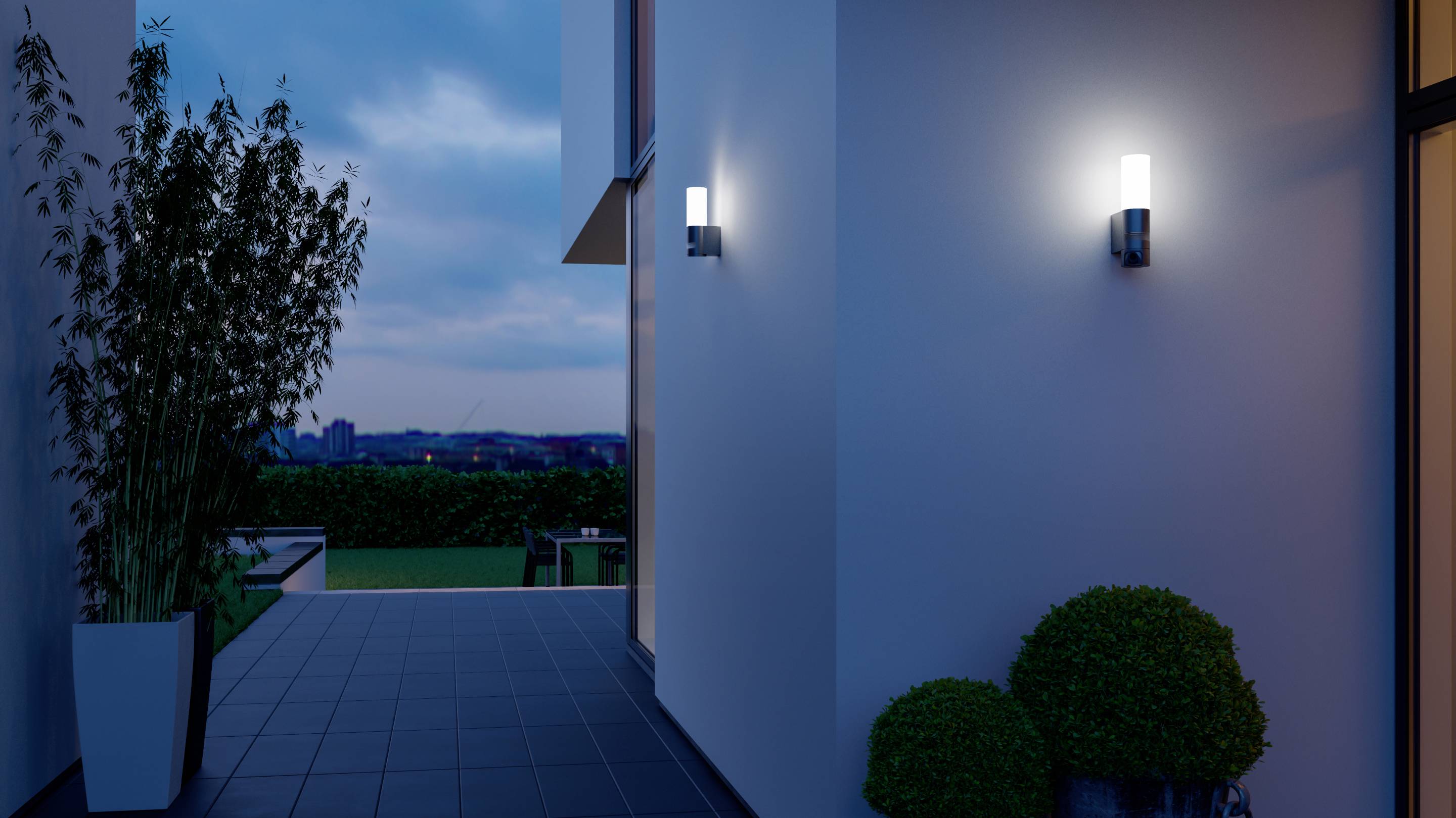 Een moderne buitenruimte bij schemering met verlichtende wandlampen aan een gebouw, planten in potten en zicht op het landschap.