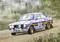 Een rallywagen rijdt over een stoffige grindweg door een bosrijk landschap. Hij draagt startnummer 2 en heeft kleurrijke sponsorlogo's.