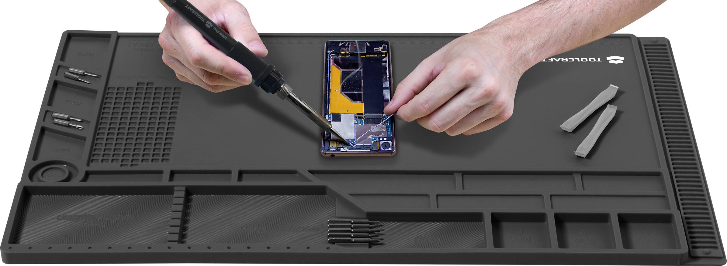 Een persoon repareert een smartphone op een zwarte anti-slip mat, omringd door schroevendraaiers en gereedschappen.