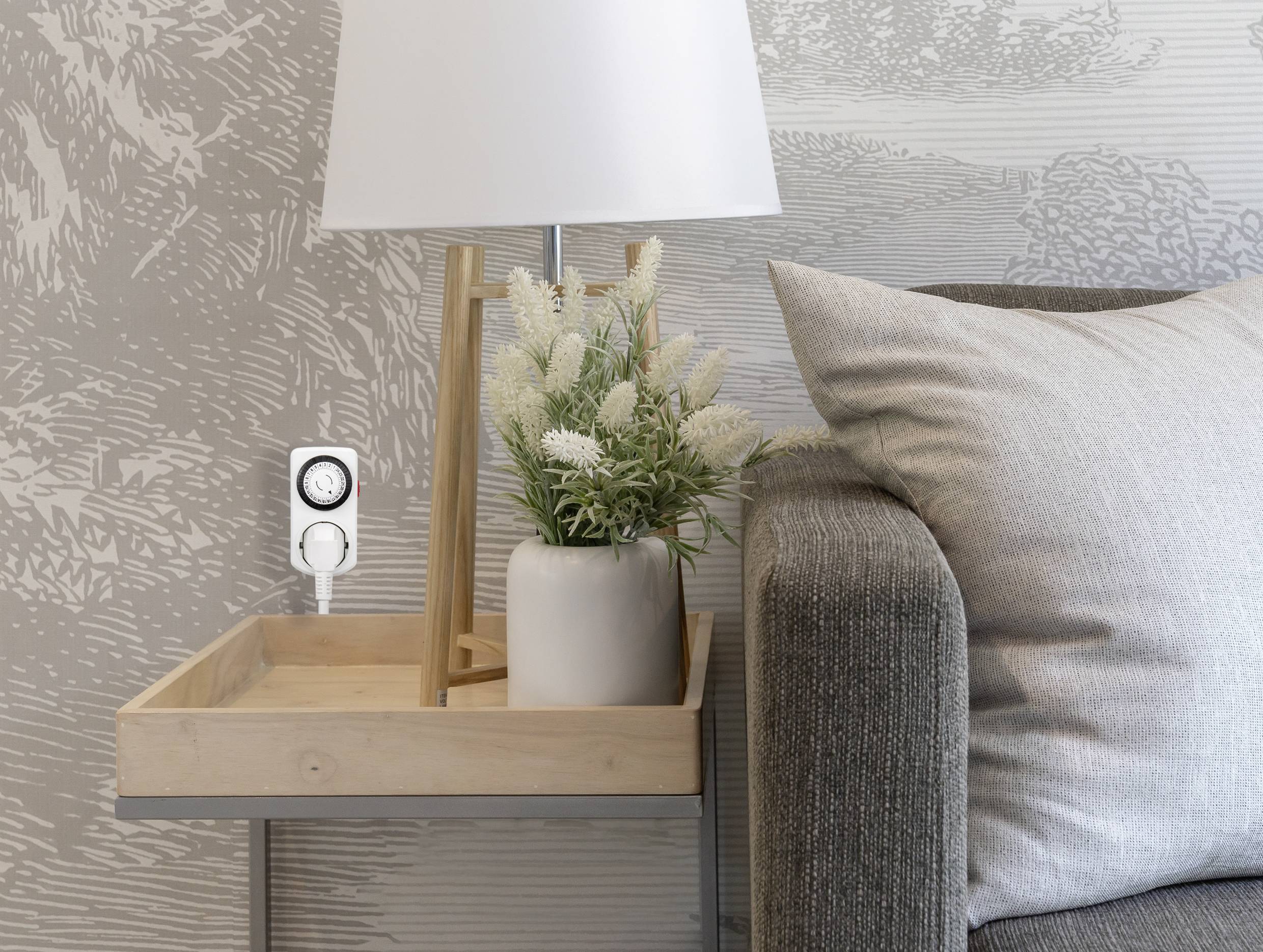 Een tafellamp en een vaas met witte bloemen staan op een houten tafel naast een grijze bank. Aan de muur hangt een tijdschakelklok in het stopcontact.