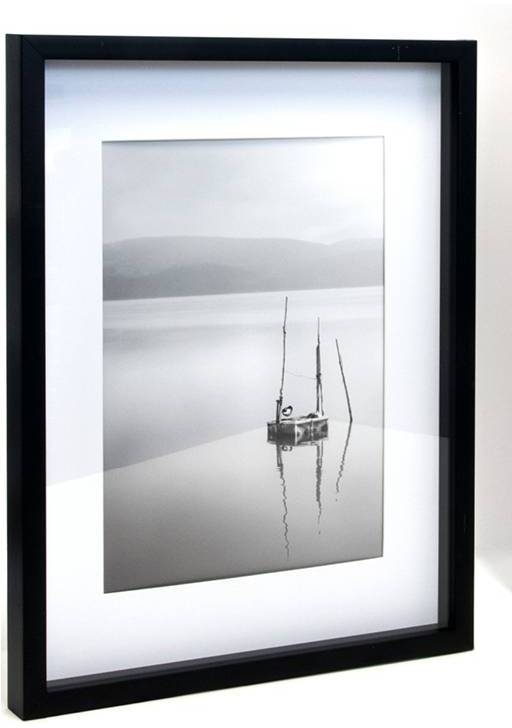 Een zwart-witfoto toont een rustige oever van een meer met bergen op de achtergrond en een kleine boot met zeilen op de voorgrond.