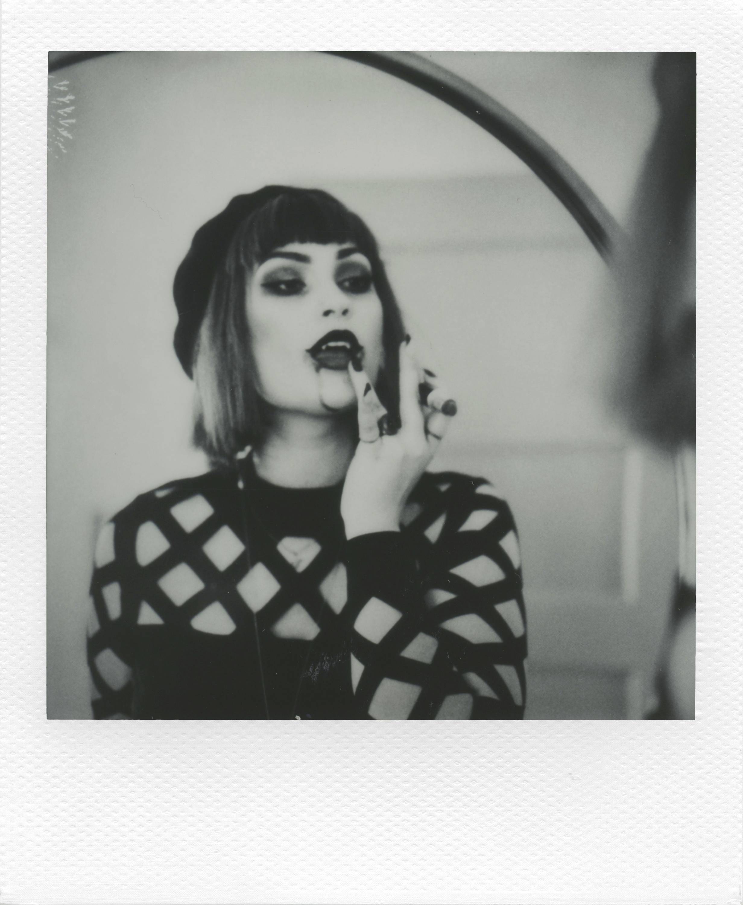 Een persoon draagt lippenstift voor een spiegel, draagt een nettrui en een muts. De opname is in zwart-wit.