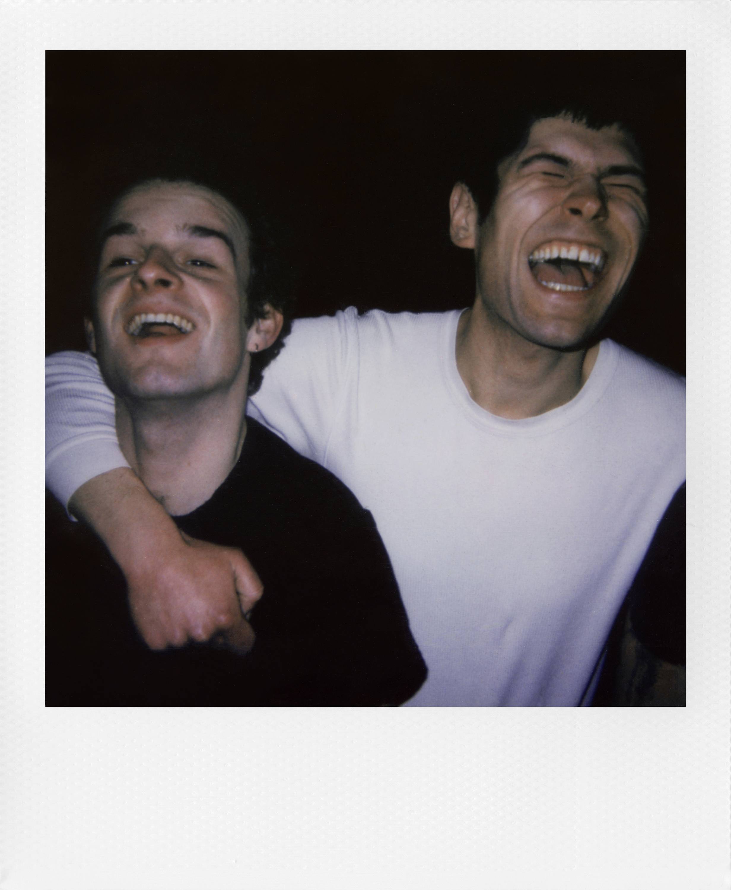 Twee personen lachen en omarmen elkaar, de ene draagt een zwart, de ander een wit bovenkleed. Ze zien er blij en ontspannen uit.