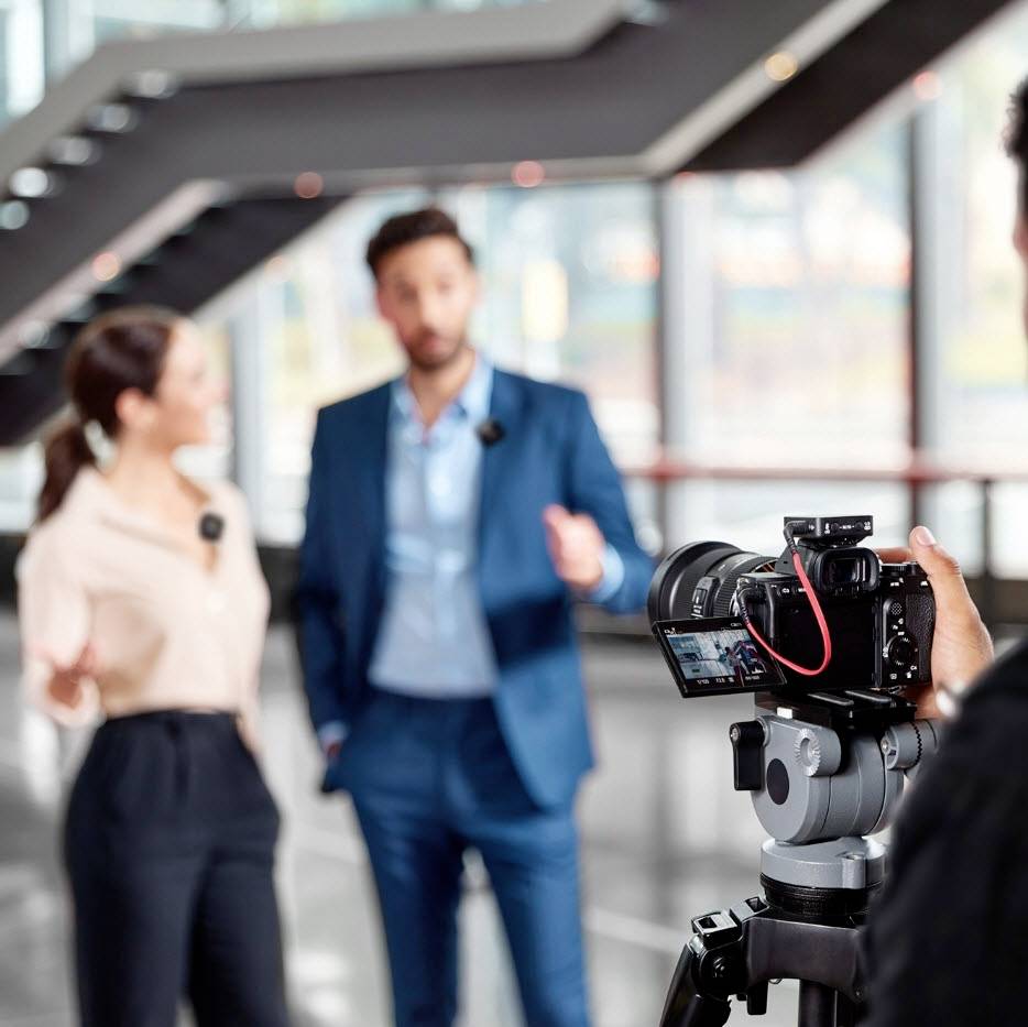 Een vrouw en een man staan voor een camera in een modern gebouw, blijkbaar in een gesprek of tijdens een interview.