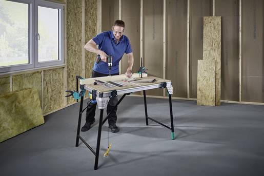 Een man met veiligheidsbril en blauwe werkkleding gebruikt een boormachine aan een tafel in een onafgewerkte ruimte met houten wandbekleding.